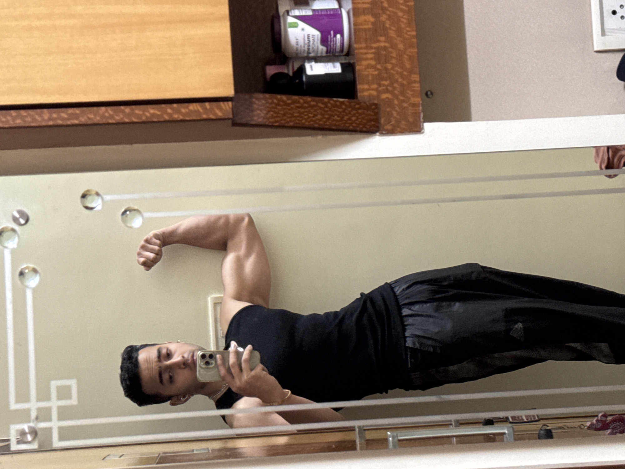 A man is taking a mirror selfie. He is wearing a black tank top and black pants. He is flexing his right bicep. In the background, there is a cabinet with bottles on top of it and a power outlet. There is no contextual evidence to suggest the location is Kolkata, India.