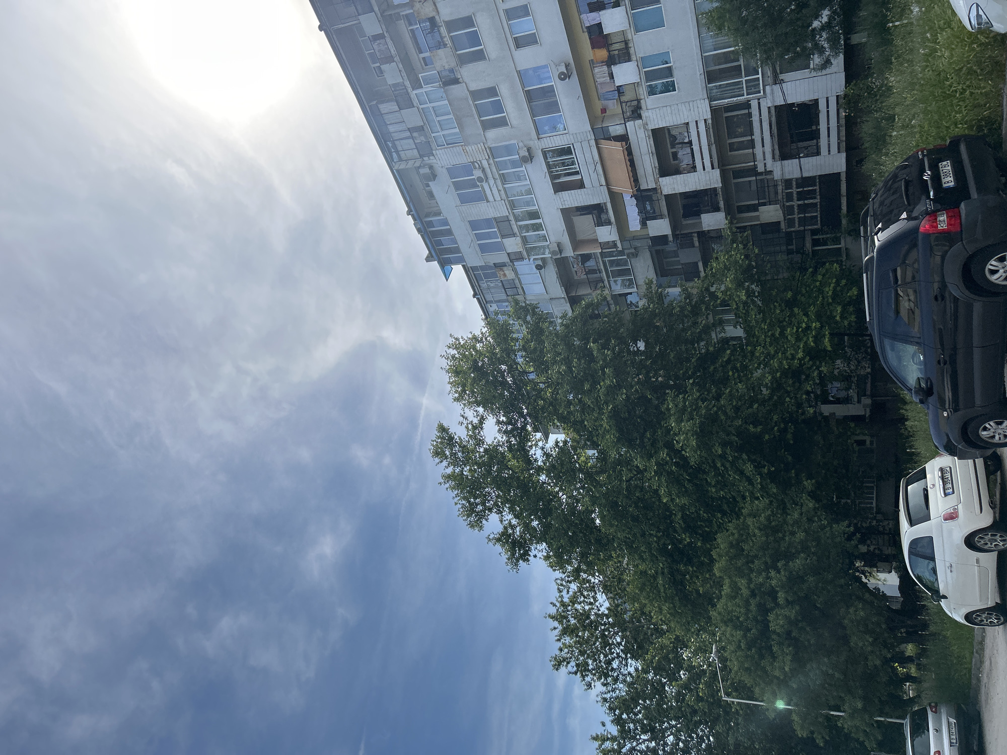 The scene depicts an urban residential environment in Varna, Bulgaria. A multi-story, light-colored apartment building dominates the right side of the frame, featuring numerous windows and balconies. Many balconies are enclosed with glass, while others are open, with some displaying beige or light blue awnings. Clothes are visibly drying on several open balconies, and multiple white air conditioning units are mounted on the exterior walls.
The left and central-left portions of the image are densely covered by large, leafy green trees, partially obscuring the lower and middle sections of the building. Below the trees and adjacent to the building, areas of unkempt green grass and weeds are visible.
Three cars are parked on the left side of the scene. From left to right: a partially visible silver hatchback (likely a Volkswagen) with Bulgarian license plate B 8414 PP; a white two-door Fiat 500 hatchback with Bulgarian license plate B 3541 BP; and a black Hyundai Tucson SUV with Bulgarian license plate B 3887 BC. A grey street light pole with a luminaire is positioned on the far left, exhibiting a green lens flare.
The sky is a bright blue with scattered thin, white clouds. The sun is intensely bright in the upper right quadrant, causing significant overexposure and glare. No people are visible in the scene. The overall lighting suggests bright daylight conditions.
