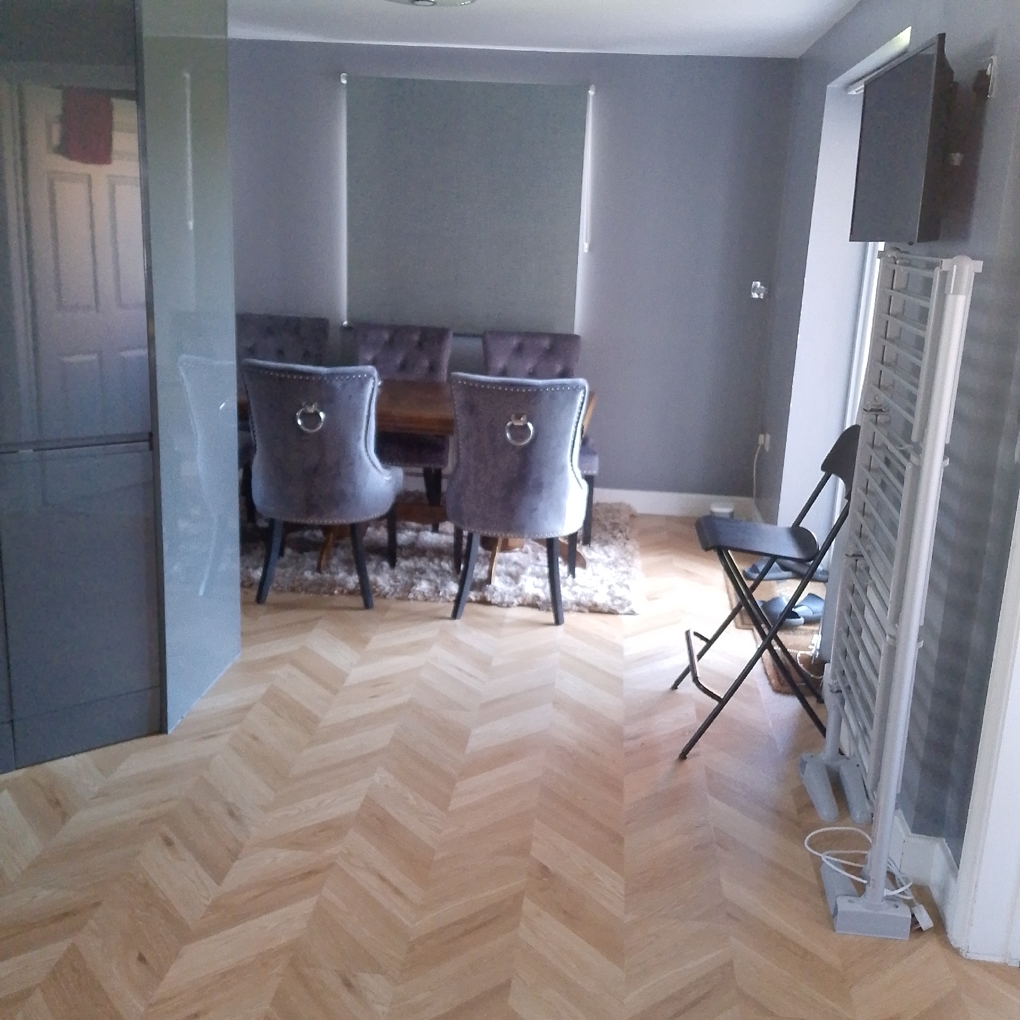 The image shows a residential interior, likely a dining room, in Corby, United Kingdom. The room is decorated in shades of gray and light wood. A dining table is surrounded by upholstered chairs with studded trim and ring pulls. A gray area rug sits beneath the table. The floor is light wood in a herringbone pattern. In the foreground, there is a gray kitchen unit to the left. To the right is a folding chair and a white electric radiator. A flat-screen TV is mounted on the wall. The blinds are closed. There are no people visible in the shot.