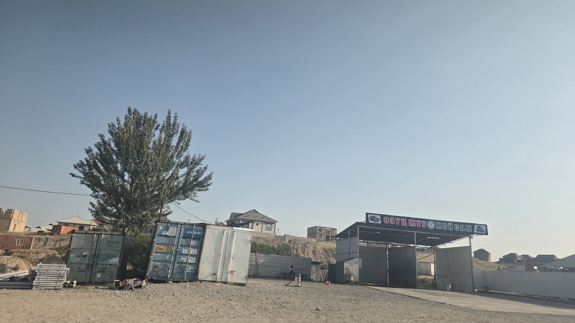 In Baş-Bulak, Kyrgyzstan, on a clear, bright day, an outdoor scene unfolds. Dominating the left side of the frame is a large, leafy tree, its branches spread wide. To its right stand several stacked shipping containers, some appearing weathered and others painted white. In front of these containers, a pile of gray pipes is neatly arranged. Further into the scene, a structure with an awning and open bays suggests a car wash, with a sign above it displaying Cyrillic text and an image of a car. The ground is covered with gravel. In the background, houses and partially constructed buildings are visible on a sloping hillside under a vast, pale blue sky. One person is faintly visible near the car wash structure.