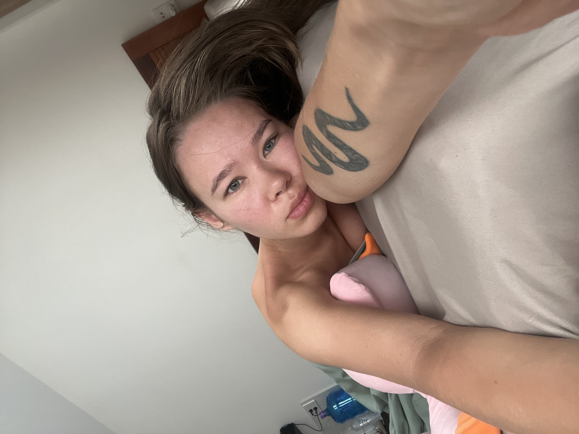 A young woman is lying in bed. The woman has light skin, brown hair, and blue eyes. She has a tattoo of a snake on her right arm. She's holding a pink stuffed animal. There is a water bottle and some other objects on the floor next to the bed. The location is likely indoors, potentially in a residence in Nam Nha Trang Ward, Vietnam, given the implied setting of a bed and domestic objects.