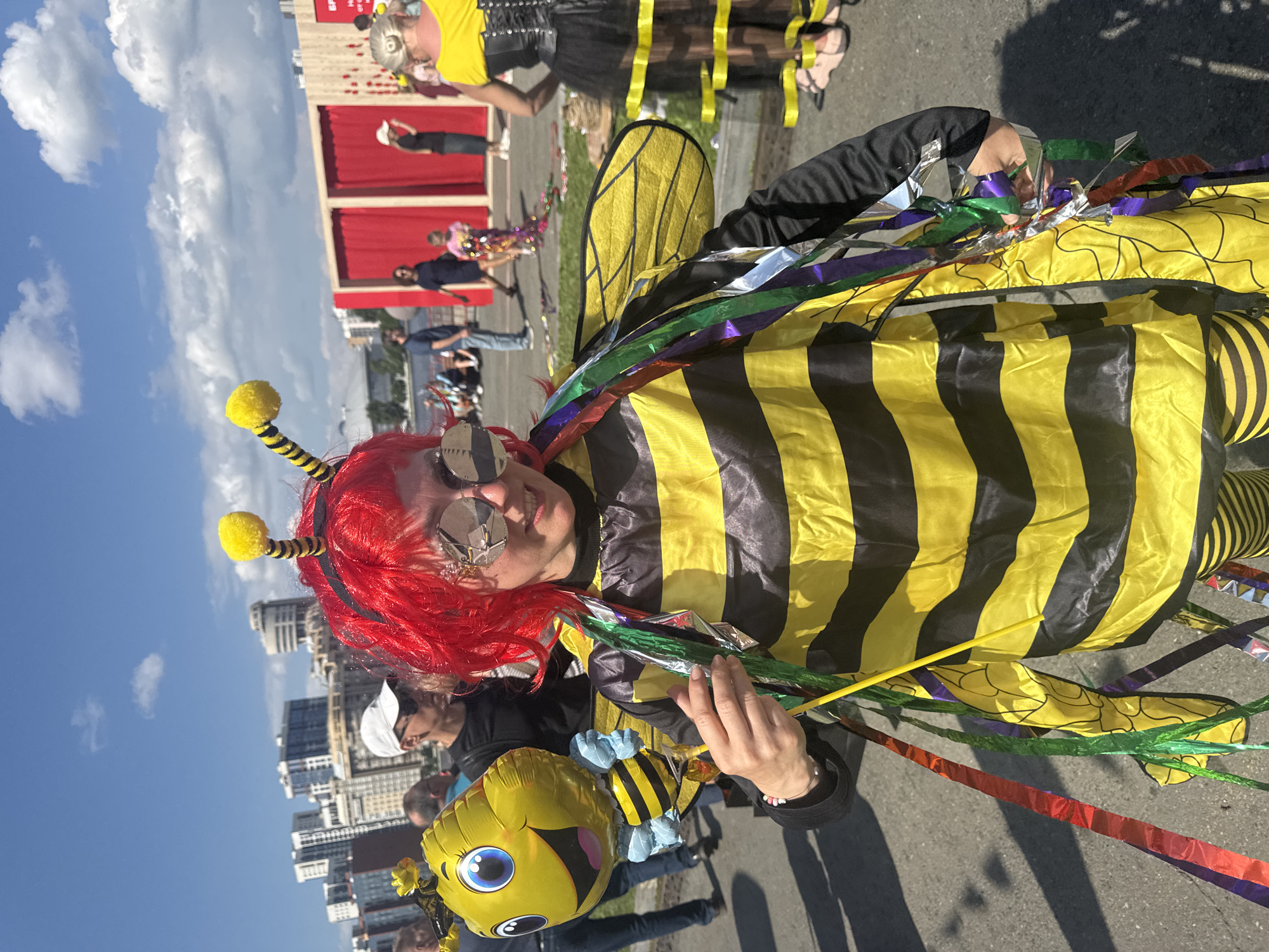 A person in a bee costume, complete with red wig and oversized, reflective sunglasses, stands holding a bee-shaped balloon. The costume features black and yellow stripes, wings, and streamers. The scene appears to be an outdoor event, possibly a festival or parade, with a crowd of people in the background. The cityscape behind them suggests an urban environment with modern buildings. The event is located in Yekaterinburg, Russia, as indicated by the text visible on the red curtains in the background, which reads "ЕКБ" (EKV, an abbreviation for Yekaterinburg) and "HORECA EXPO". The weather is sunny with a few clouds in the sky.