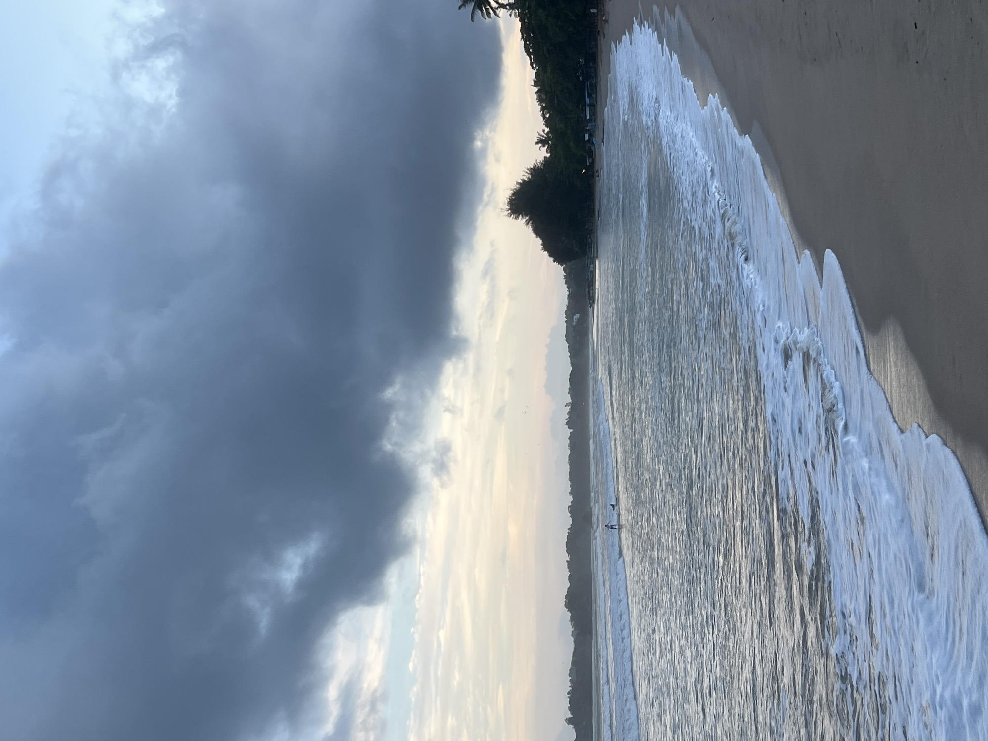 A wide shot captures a serene beach scene in Peleana, Sri Lanka, under a dramatic, overcast sky. The foreground features a sandy beach with gentle waves lapping at the shore, leaving a foamy white trail. In the mid-ground, the ocean stretches out, with two figures on paddleboards visible in the distance. A line of trees and vegetation forms a dense backdrop along the coastline, with a few small structures peeking through the foliage. The overall impression is one of natural beauty and tranquility, with a hint of impending weather change due to the dark, heavy clouds overhead.