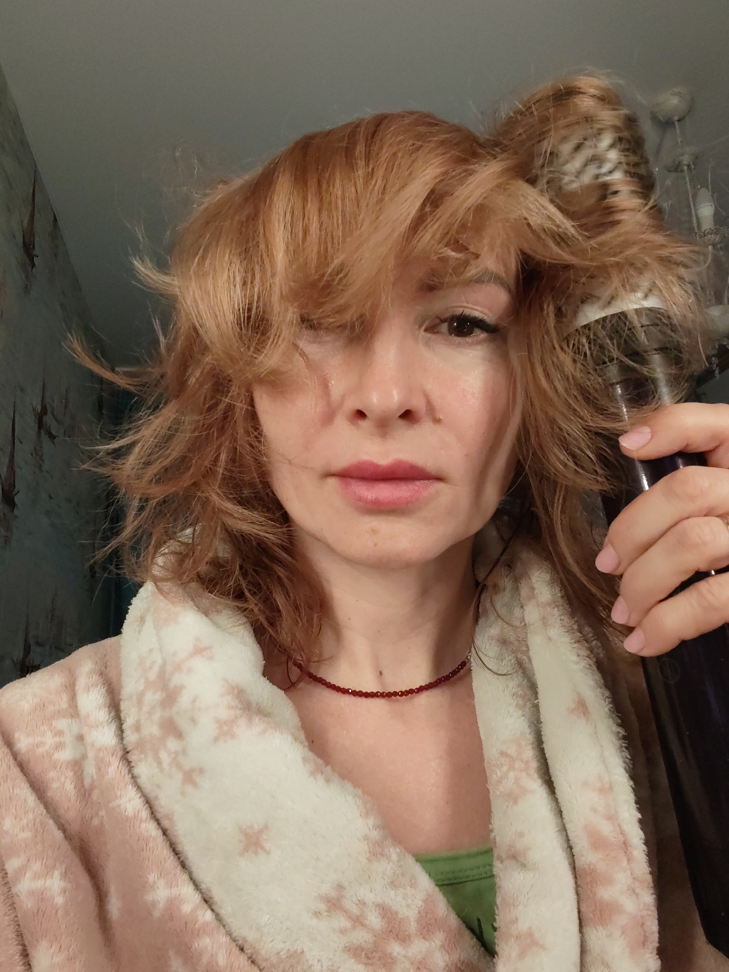 The image depicts a female individual in an indoor setting within the city of Chaikovsky, Russia. The subject has light reddish-brown, wavy hair, parted on the left, with strands falling across her forehead and partially obscuring her left eye. Her right eye is visible, with a dark winged eyeliner. Her lips are slightly parted, exhibiting a natural pink hue. She wears a light pink, plush robe with a white collar and white snowflake patterns. A thin, red beaded necklace is visible around her neck, and a green garment is partially visible beneath the robe. Her right hand, displaying light pink manicured nails and a ring on the ring finger, holds a dark, cylindrical object, possibly a hair styling tool, positioned near the right side of her head, intertwined with her hair. The background consists of a dark teal or greyish-blue wall with a faint, repeating pattern, likely wallpaper. In the upper right corner, a portion of a white light fixture with multiple elements is discernible against a lighter ceiling. The scene appears to be an interior, possibly a domestic space.