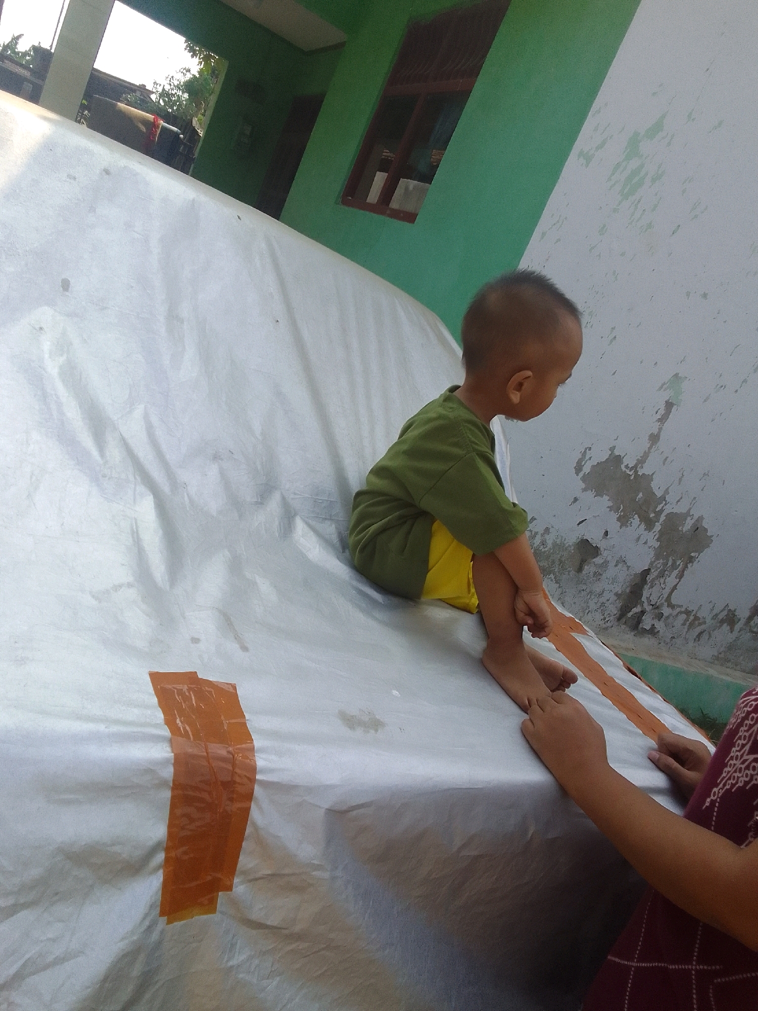 In a residential setting in Karangcegak, Indonesia, a young child, appearing to be a toddler, sits on a large, white, tarp-covered surface. The child is wearing a green t-shirt and yellow shorts and is looking to the right. Two strips of orange tape are affixed vertically on the tarp. To the right of the child, a pair of adult hands, belonging to an individual wearing a dark patterned garment, are visible, with one hand touching the child's foot. The background includes a portion of a building with a green wall and a window with brown trim. The overall scene suggests a casual outdoor activity.