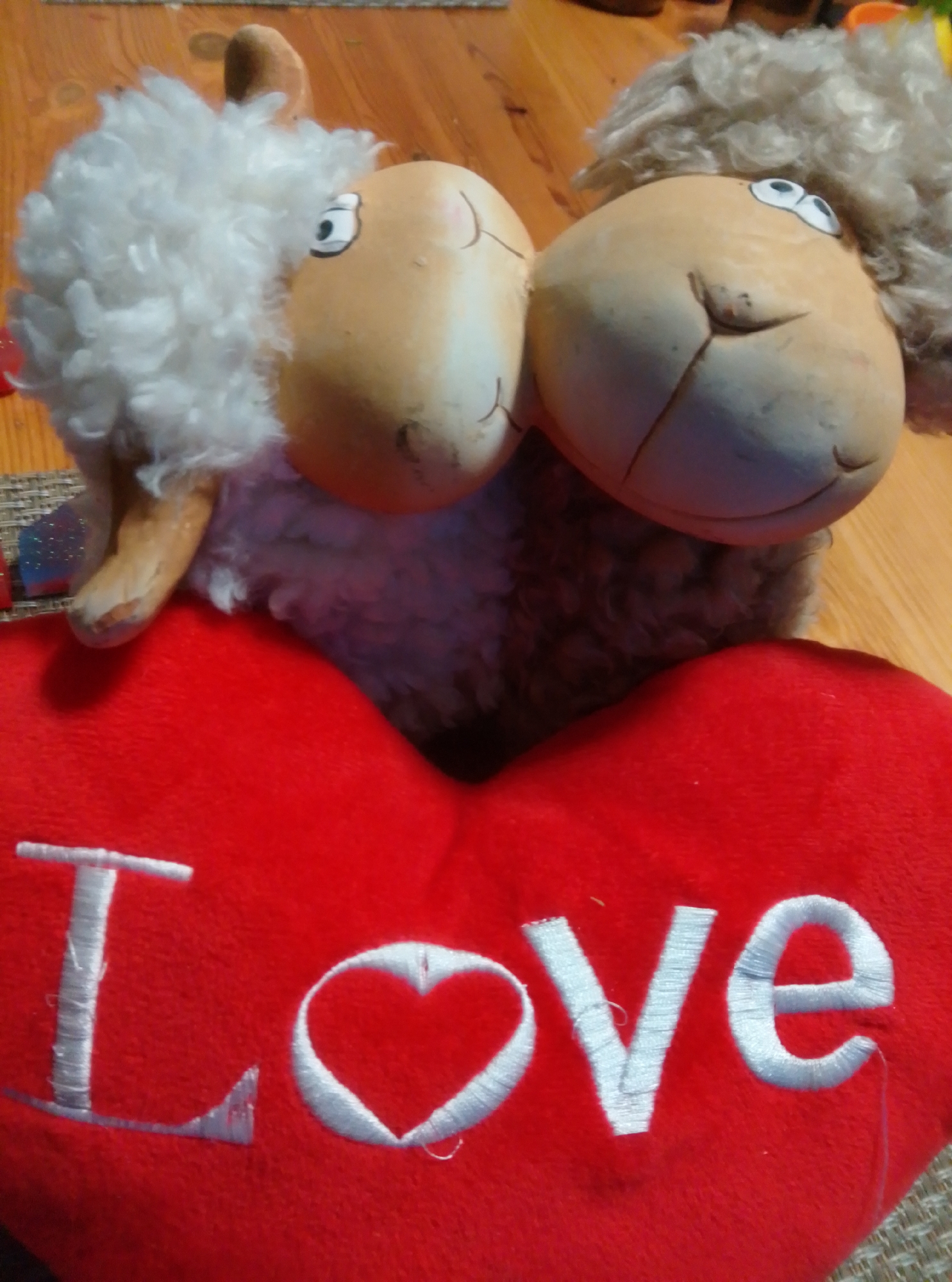 The image shows a close-up of two stuffed sheep figures positioned on a red heart-shaped pillow. The sheep have soft, fluffy wool and cartoonish faces with large eyes and simple smiles. The red pillow is embroidered with the word "Love" in white thread. The background appears to be a wooden surface, possibly a table, with a woven mat visible in the lower left corner. The image was taken in the city of Wedel, Germany.