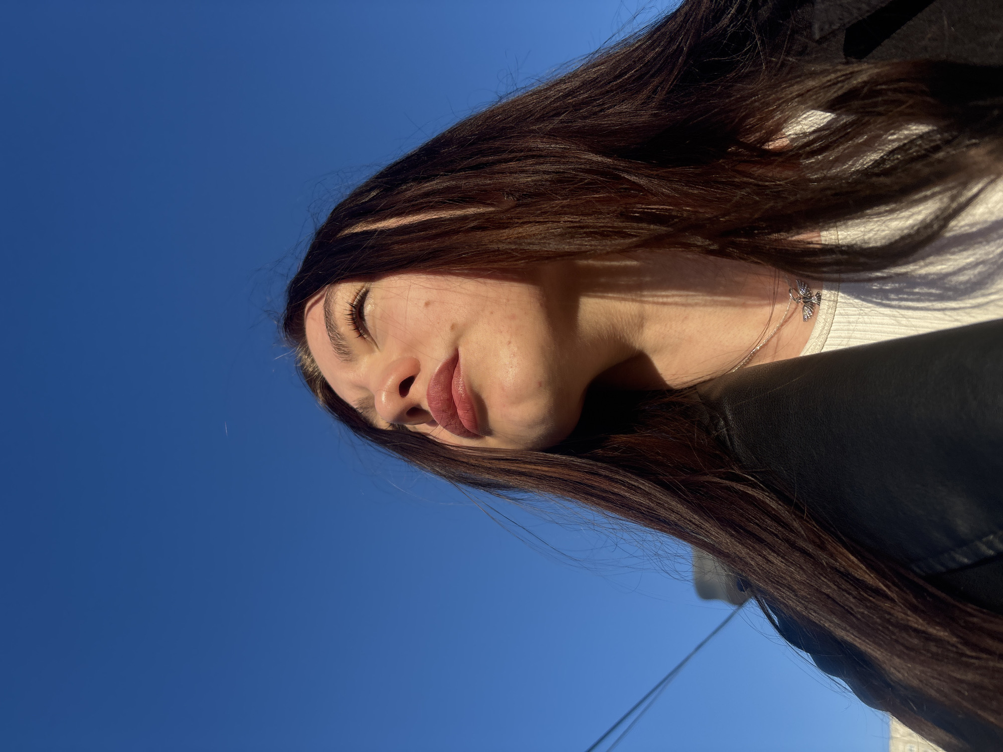 A young woman with long, dark brown hair is visible against a clear, deep blue sky. She appears to be looking slightly downwards. Her eyes are closed. Her facial features include a nose, lips with lipstick, and clear skin. She is wearing a white top and a black jacket. A delicate silver necklace with a small charm is visible around her neck. The lighting suggests it is daytime and the sun is at an angle, casting shadows on her face and hair. The image composition suggests it was taken from a low angle, looking upwards. There is no information in the image to indicate a location in Perm, Russia.