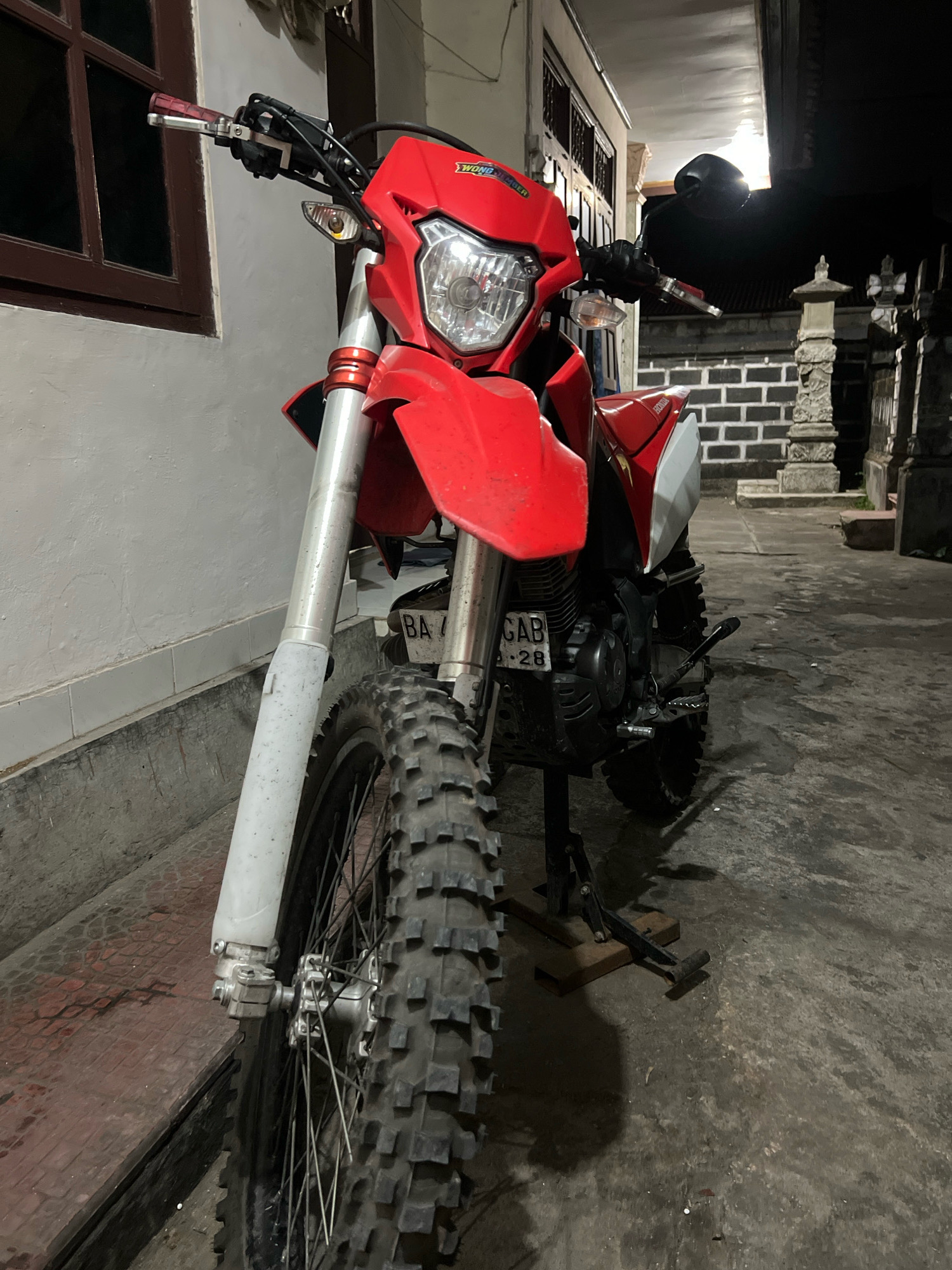 A red and white dirt bike is parked on a paved surface at night. The bike is upright, supported by a kickstand resting on wooden blocks. The bike's front tire, with knobby tread, is prominently featured. The motorcycle has a red fuel tank and fairing, with white accents on the rear. A license plate, partially visible, reads "BA / 28". In the background, a brick wall and stone pillars suggest an outdoor courtyard or alleyway. To the left, a building with a dark-framed window is visible. The lighting suggests an artificial light source casting shadows. The overall environment appears to be an urban setting in Denpasar, Indonesia.