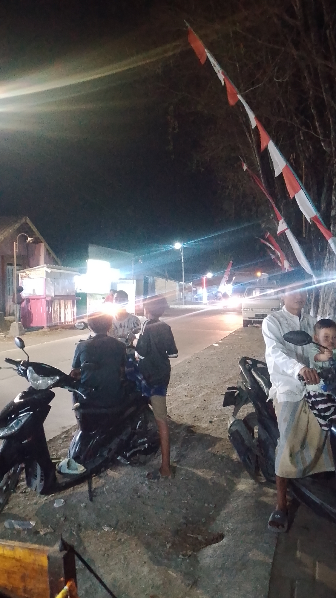At night in Karanglangu, Indonesia, several people are gathered near motorcycles on a dirt roadside. The scene is illuminated by bright artificial lights, including streetlights and vehicle headlights. To the left, a black motorcycle is parked, with three individuals in the background, two of whom are sitting on another motorcycle. One person, wearing a dark shirt, is seated, while another, in a patterned shirt, is also seated. A third individual in a dark shirt and shorts stands next to them.
Further to the right, a man in a white shirt and a light-colored sarong is seated on a motorcycle, holding a young child. Red and white flags are strung along the side of the road, attached to trees and poles. In the background, a road with passing vehicles is visible, along with several buildings, some of which appear to be small shops or stalls. The overall atmosphere is casual and communal, suggesting a gathering or a stop along a route.
