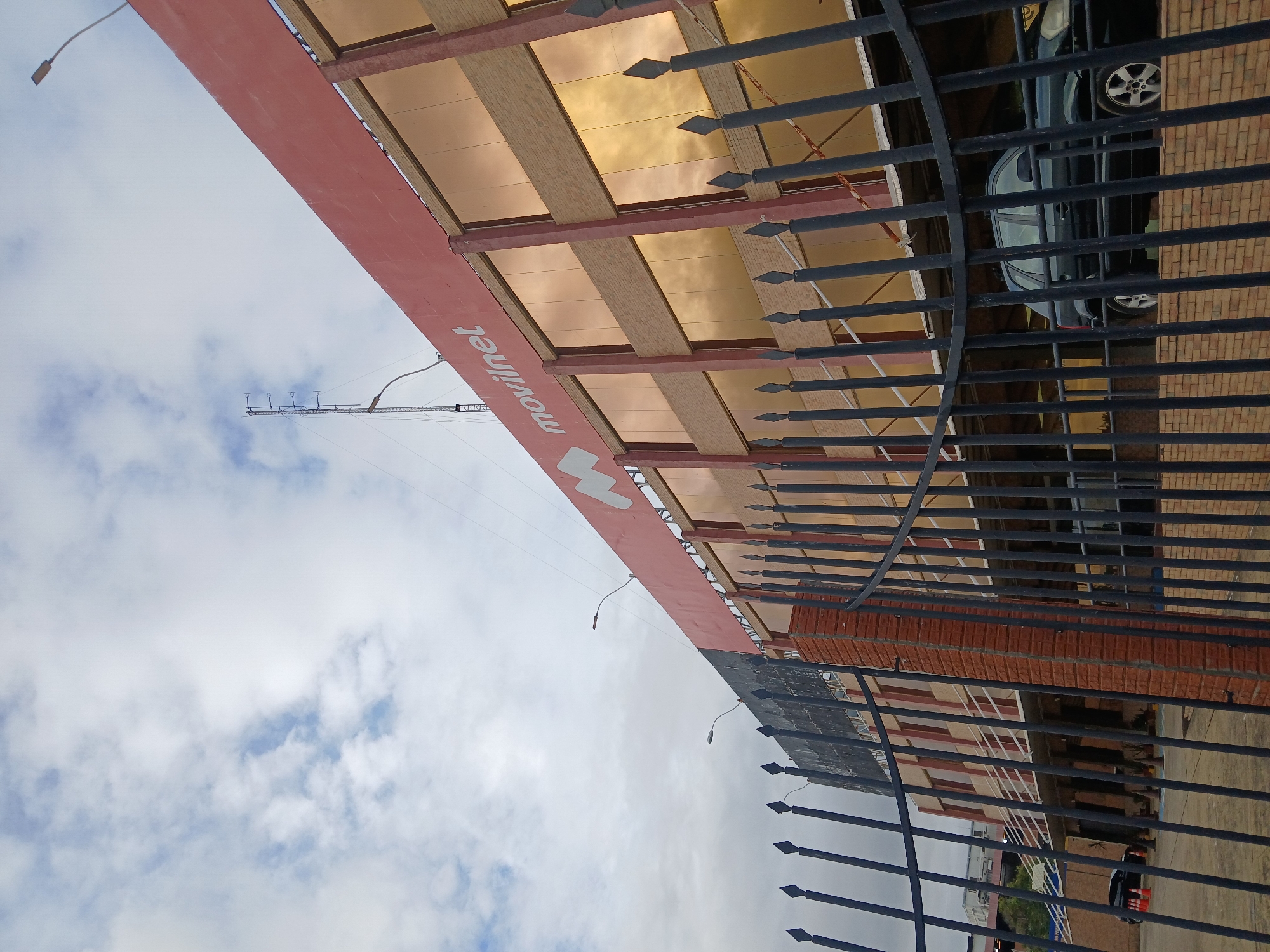 A low-angle exterior view depicts a multi-story Movilnet telecommunications building in Ciudad Guayana, Venezuela, under a partially cloudy sky. The building features a prominent red upper facade displaying the "movilnet" name and its stylized 'M' logo in white. Below this, the structure presents several floors with reflective, golden-tinted windows separated by reddish-brown vertical columns and horizontal brick-patterned sections. A communication tower equipped with multiple antennas is visible extending from the building's roof. Streetlights are mounted on the building's exterior. In the immediate foreground, a dark metal fence with spiked finials, built upon a low brick wall, partially obstructs the lower portion of the building. Through the fence, a dark-colored SUV is parked on an elevated section of the building's access area, and several orange traffic cones are arranged on the paved ground below. A second dark vehicle is partially visible further back on the left. No individuals are present in the scene.