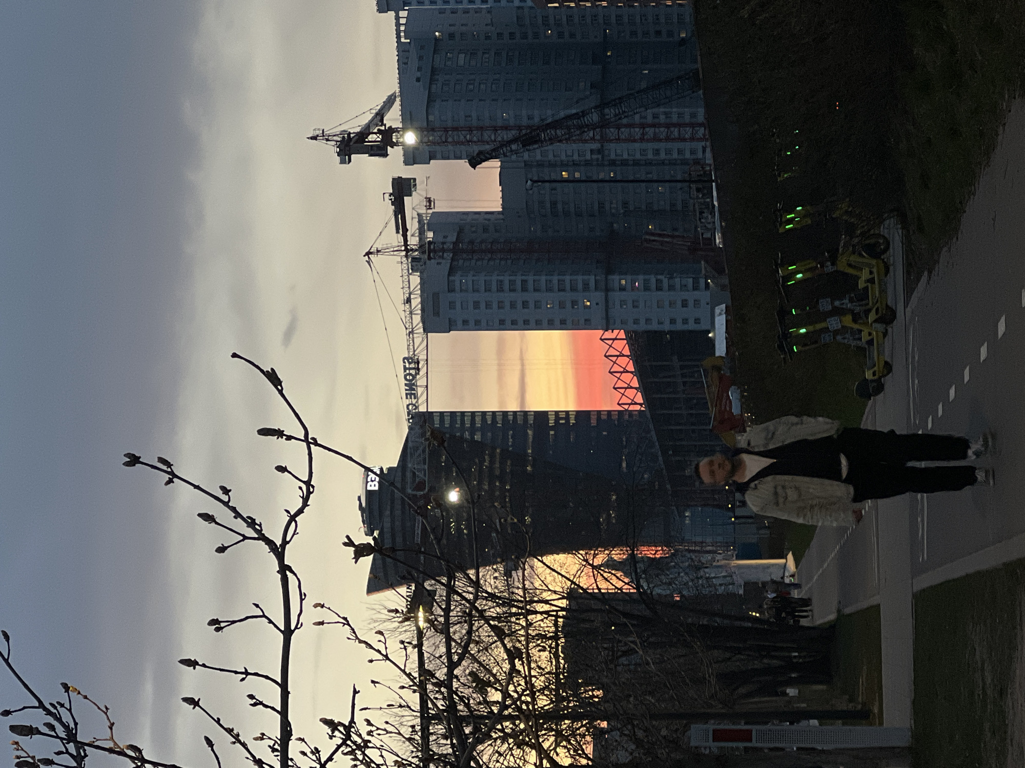 The image shows an urban landscape in Moscow, Russia, at sunset. Tall modern buildings are visible in the background, some under construction with cranes. A person is standing on a sidewalk, partially obscured by trees. A row of parked electric scooters is also present. The sky exhibits a gradient of colors ranging from gray to orange, reflecting the setting sun.