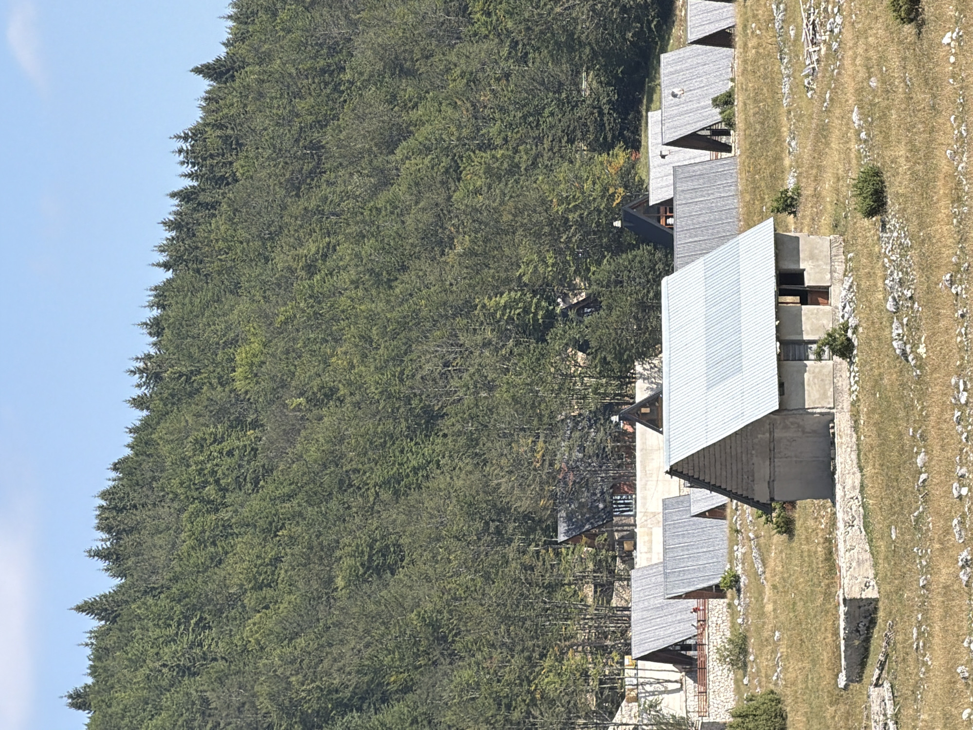 In Virak, Montenegro, a cluster of rustic, A-frame cabins with corrugated metal roofs dot a grassy hillside. The cabins are primarily constructed from wood and stone, with some appearing newer and more finished than others. In the foreground, a single, simpler cabin with concrete walls and a metal roof stands prominently. The surrounding landscape is characterized by dry, golden grass, scattered rocks, and low-lying bushes. Behind the cabins, a dense forest of coniferous trees rises to meet a clear blue sky. The scene suggests a remote, natural setting, possibly a mountain village or a tourist resort.