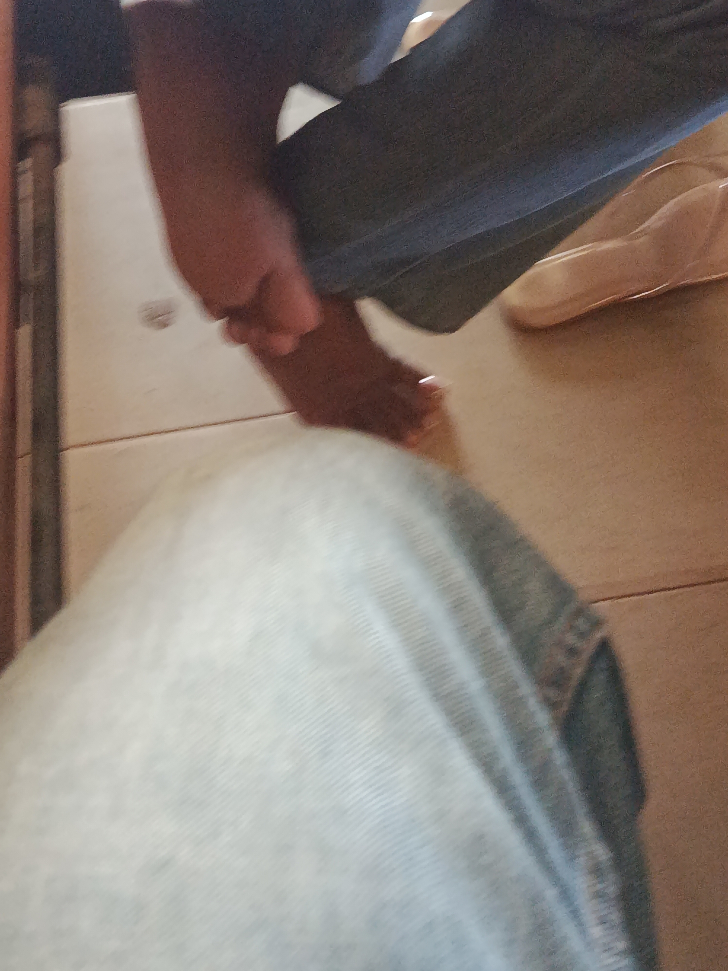 In an indoor setting with tiled flooring, a person's left foot is visible, resting on the floor. The foot is being held by a hand. The person is wearing blue jeans. A light-colored shoe is also visible on the floor. The background consists of a wall.