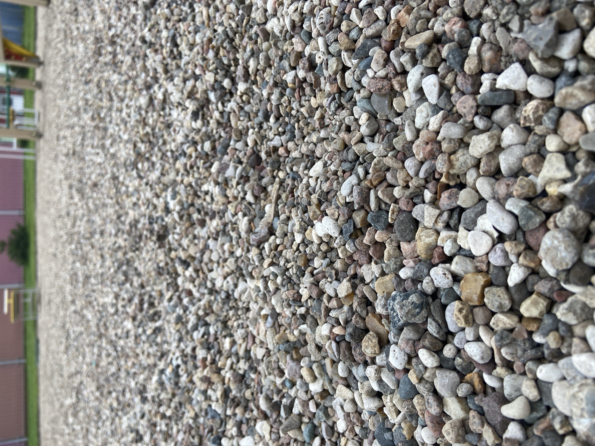 The image is a close-up, low-angle shot of a gravel surface, likely on a playground or park. The gravel consists of small, multi-colored stones, predominantly in shades of gray, white, brown, and some with reddish hues. The stones are varied in shape and size, creating a textured and uneven ground. In the background, slightly out of focus, is a playground structure with some yellow and green elements, and a pink building facade. The environment appears to be outdoors, with green grass visible in the mid-ground. The scene is static, with no people or animals present. The focus is entirely on the gravel, emphasizing its granular nature and the play of light and shadow on its surface. No specific location in Balashikha, Russia can be determined from the image itself; it is a generic outdoor scene.