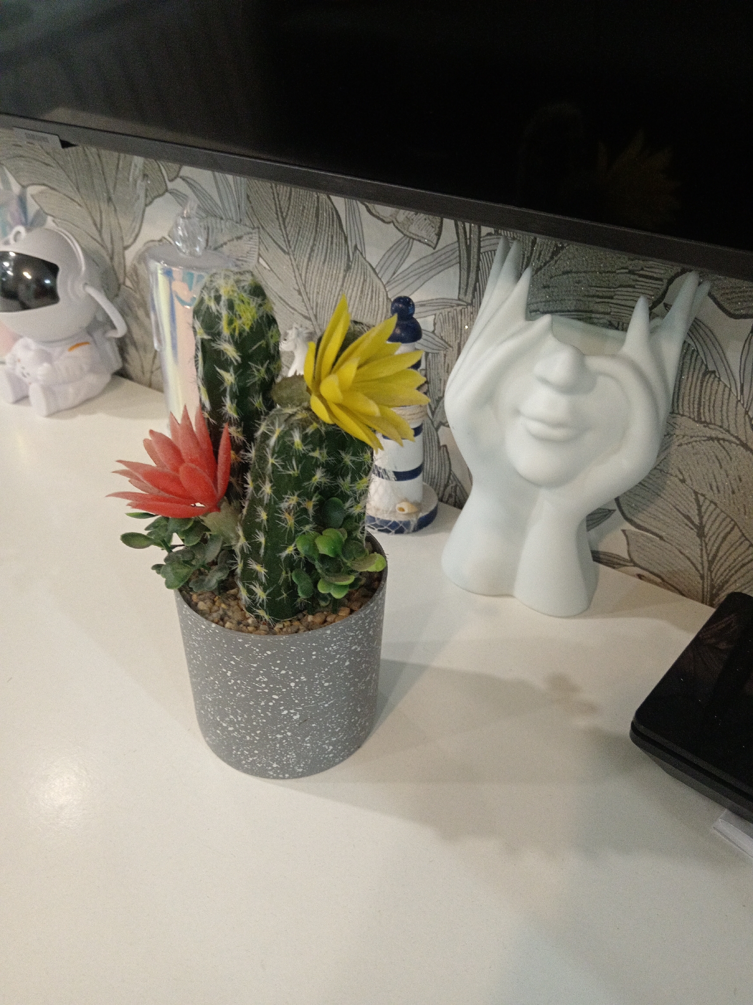 A close-up, overhead shot shows a small potted artificial cactus with red and yellow flowers on a white, speckled surface. The cactus is in a gray speckled pot with pebbles on top. Behind the cactus, to the left, is a white astronaut figurine. To the right of the cactus, a white sculpture of a face with hands covering its eyes is visible. In the background, a wall with a leaf pattern wallpaper is seen. A black flat-screen TV is mounted above the wallpaper, reflecting a dim light. A black electronic device, possibly a phone or tablet, is on the right side of the surface. The scene appears to be indoors, on a shelf or table, with no indication of a specific location in Belogorsk, Russia, or any specific actions or interactions taking place.