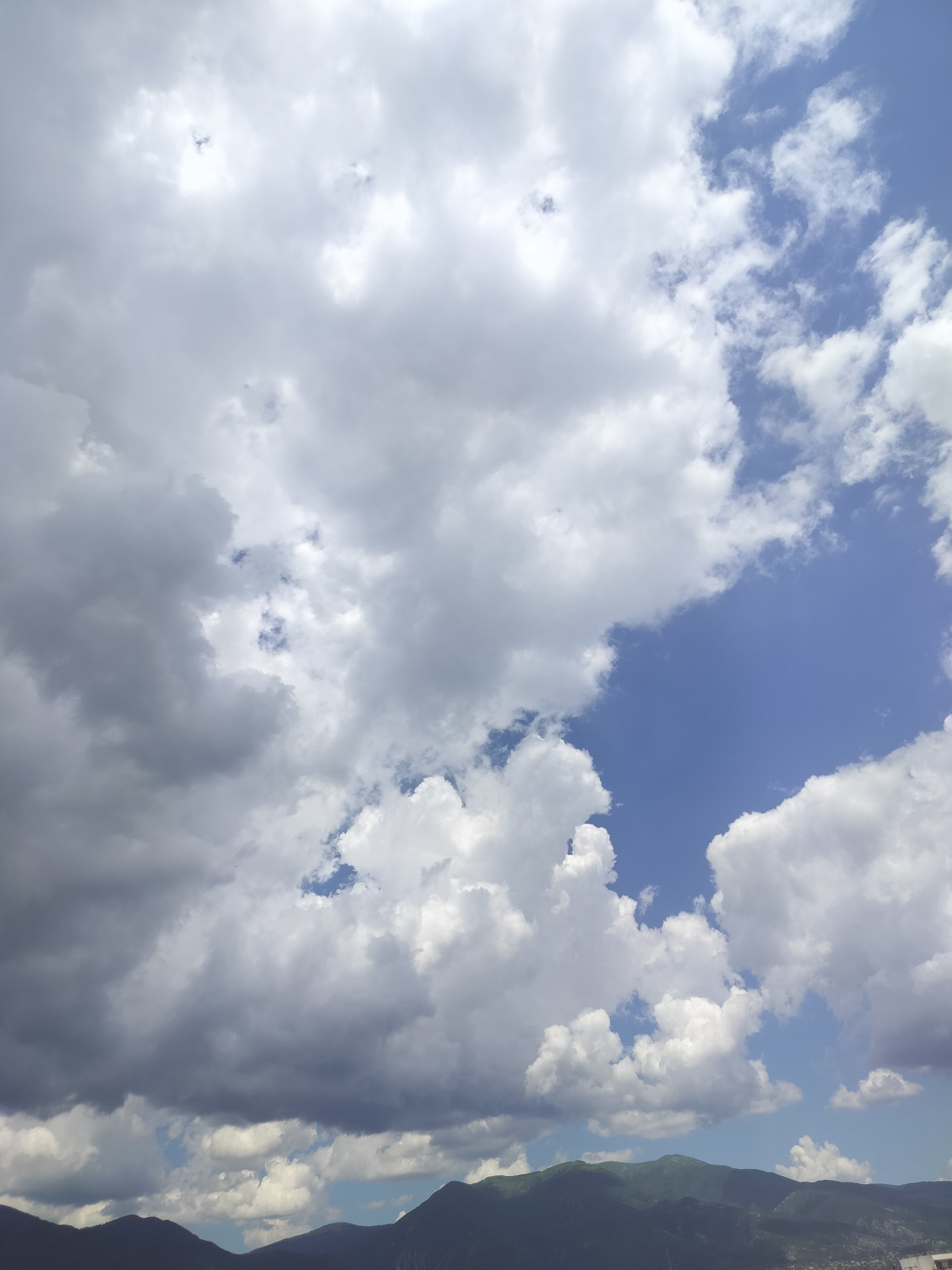 The image depicts a sky filled with a mix of white and gray clouds, with patches of blue sky visible. In the lower portion of the image, a dark, undulating mountain range stretches across the horizon. The scene appears to be a landscape view, likely taken from an elevated position within or near the city of Gorno Sahrane, Bulgaria, given the mountainous terrain. No people or specific structures are clearly identifiable within the frame.