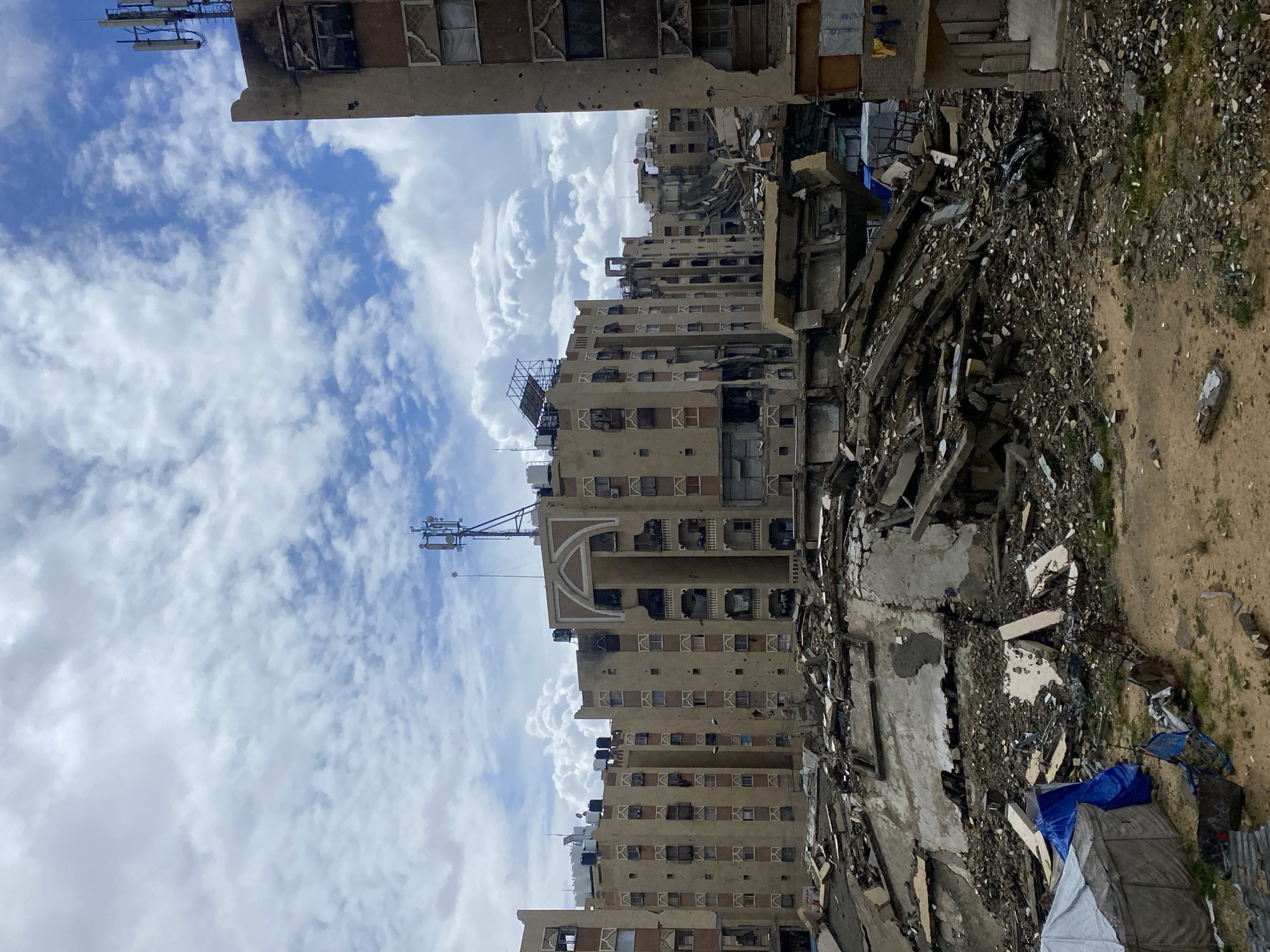 The image depicts a devastated urban landscape in Khan Yunis, Palestinian Territory, under a sky with scattered white cumulus clouds. The foreground shows a sandy-brown terrain interspersed with rubble and debris. In the lower-left, a light-colored tent is partially covered by a blue tarp, with additional blue fabric nearby. The midground is dominated by a massive mound of collapsed concrete and rebar, indicating a completely destroyed structure. To the right, the lower section of a building exhibits heavy damage, with yellow fabric hanging from its exterior.
In the background, a series of multi-story apartment buildings, approximately eight to ten floors tall, line the horizon. These structures show extensive damage, including numerous shattered windows, exposed darkened interiors, and widespread impact marks on their facades. Several buildings display charred sections, indicating fire damage. Black water tanks are visible on many rooftops. A prominent building in the center features a decorative arched element on an upper floor, also showing signs of damage. Cellular communication towers with multiple antennae are mounted on the rooftops of two buildings, one centrally located and another on the far right. The building on the far right edge of the frame is heavily damaged, with visible rebar and broken window frames, and its facade displays multiple perforations consistent with bullet or shrapnel impact. No individuals are visible in the scene.