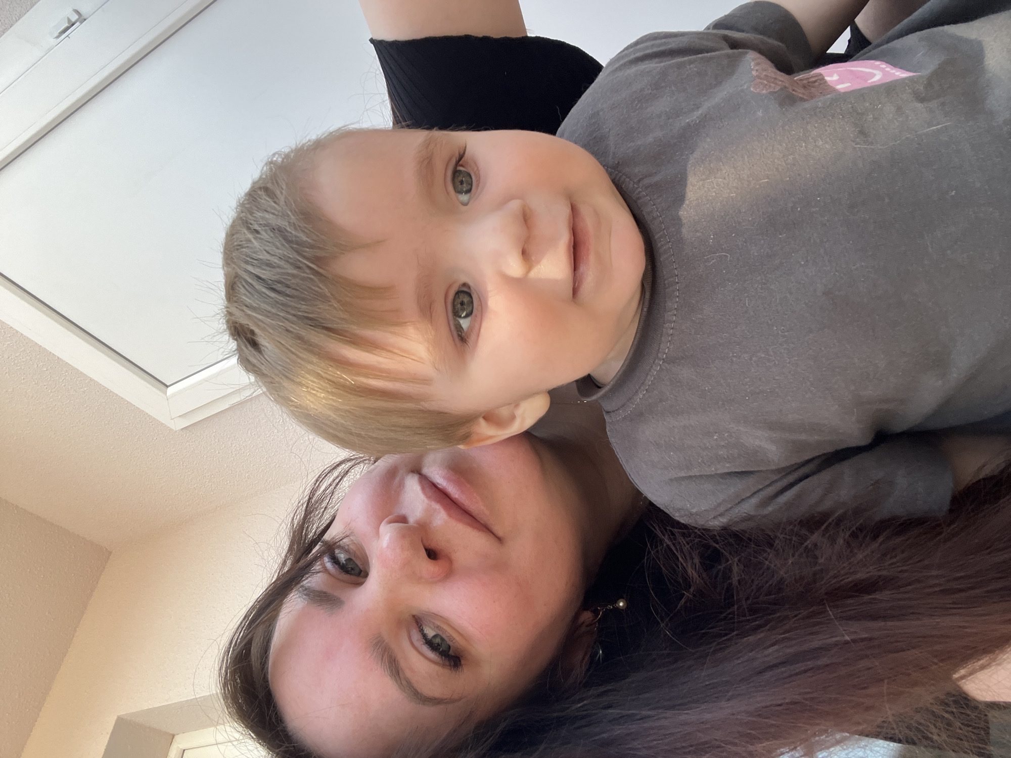 A selfie taken from a low angle shows a woman and a baby. The woman has dark hair and is wearing pearl earrings. The baby is wearing a gray shirt with a pink design on the shoulder. The background includes a white ceiling and wall. The location in Pervomaysky, Russia is not discernible from the image.