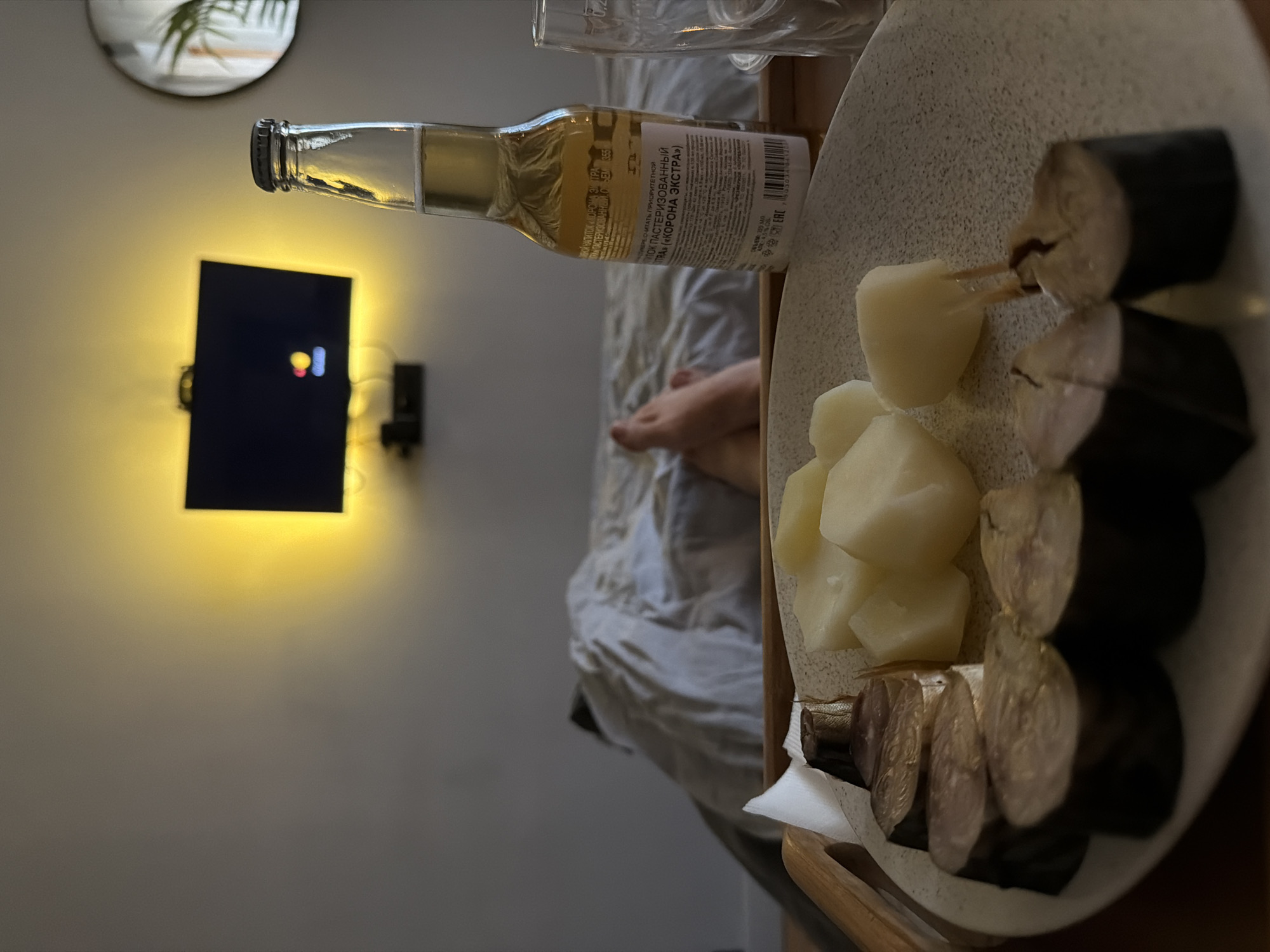 In a dimly lit room, likely a bedroom in Khimki, Russia, a meal is presented on a wooden tray. The focus is on a plate of sliced smoked fish and boiled potatoes, alongside a bottle of amber liquid, possibly beer, and a partially visible glass. In the background, a person's feet are visible under a light-colored duvet, suggesting they are resting in bed. A television screen, illuminated by a yellow backlight, is mounted on the wall above the bed, displaying a colorful icon. A round mirror with a plant is also visible on the wall. The overall impression is one of a relaxed, solitary evening meal.