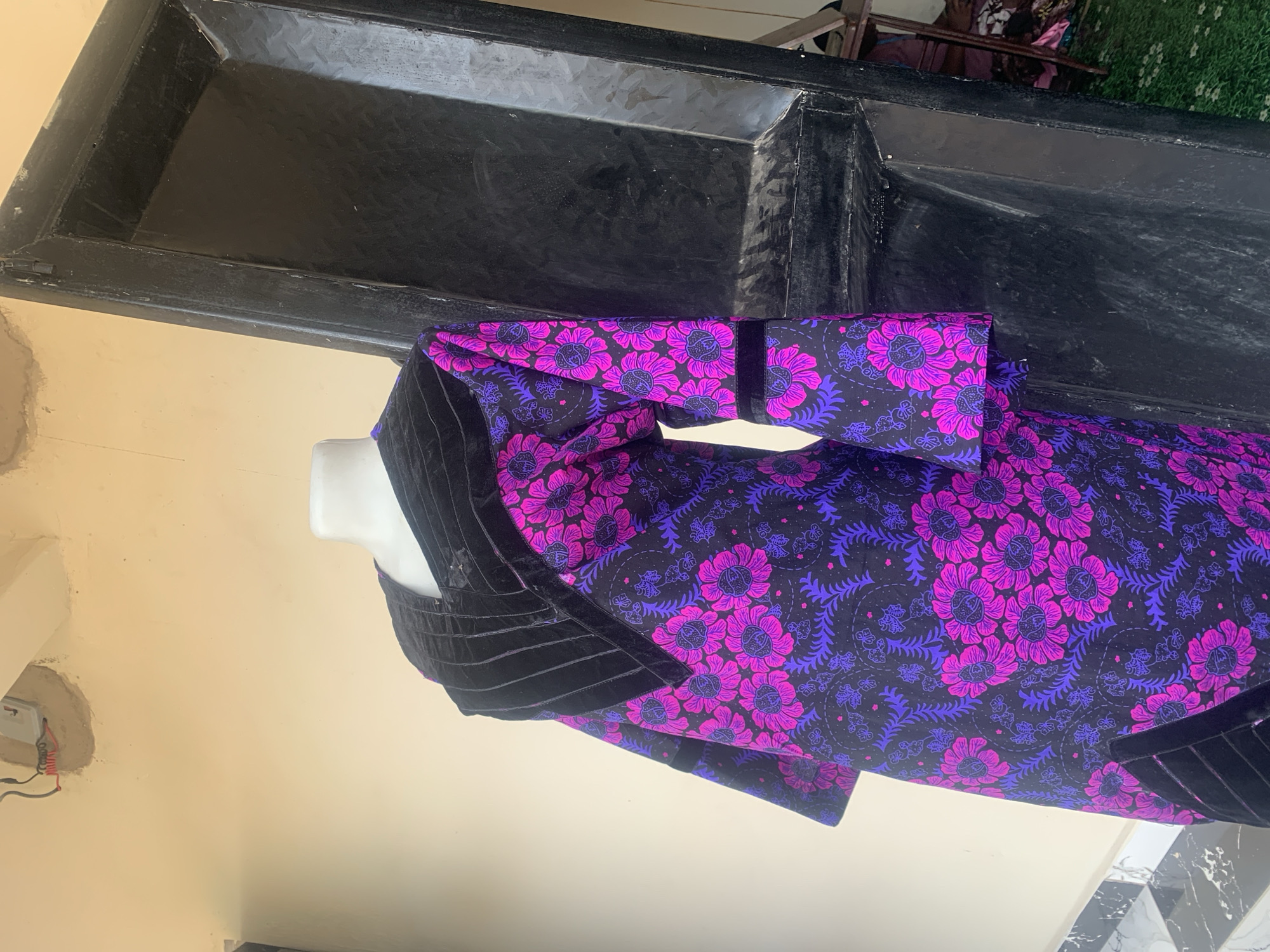 A headless mannequin, dressed in a vibrant purple and pink floral print dress with black velvet trim, stands indoors. The dress has wide sleeves and a unique collar. The background consists of a light-colored wall with an electrical box, a large black door, and some tiling on the floor. The scene appears to be in a retail or display setting, possibly a boutique in Jauro Yinu, Nigeria.