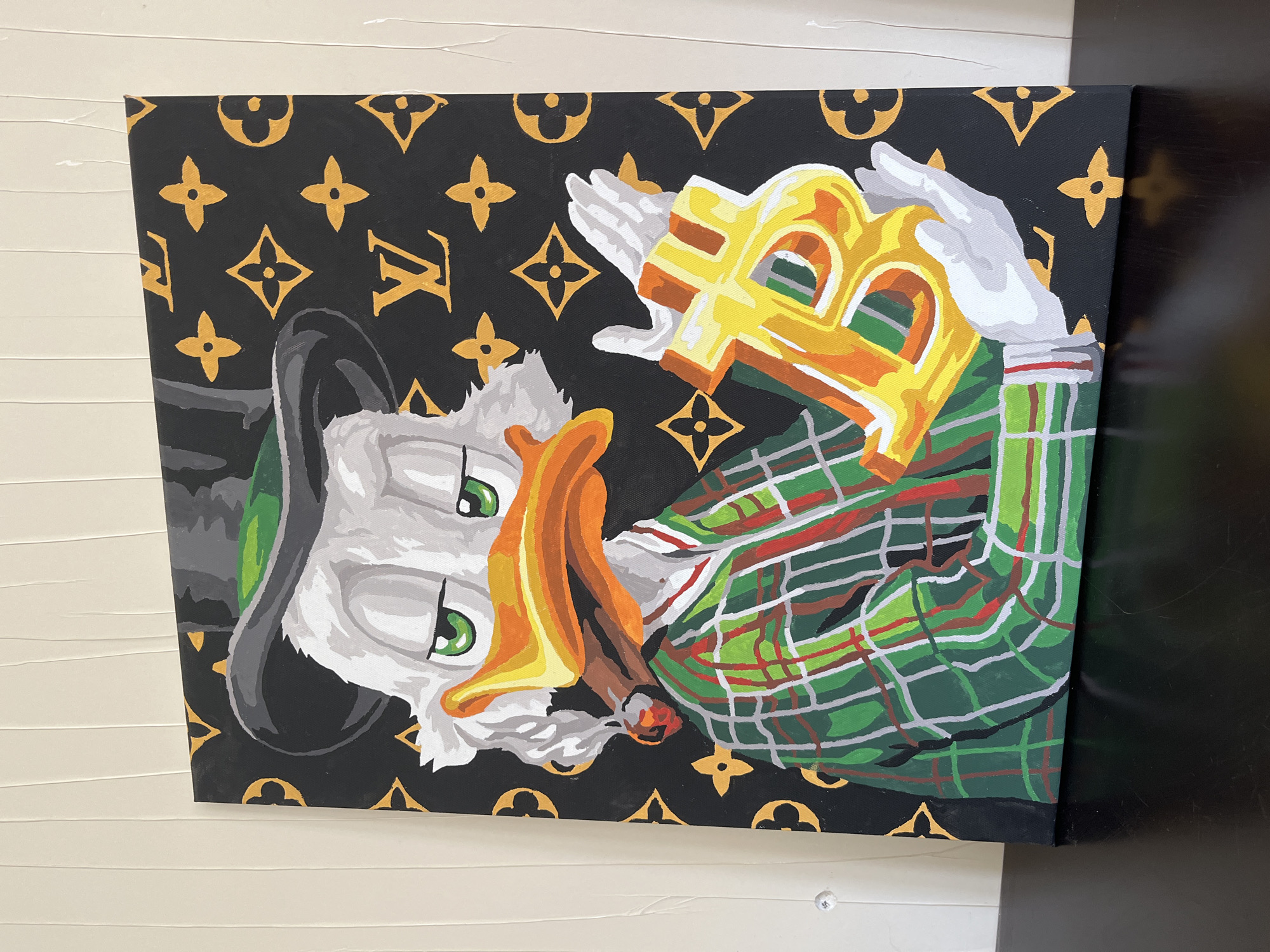 The image shows a painting of Scrooge McDuck on a black background with gold Louis Vuitton logos. He is wearing a green plaid jacket and holding a large gold "B" in his hands. He has a cigar in his mouth. The painting is leaning against a white wall with horizontal grooves and a black surface on the right side. There is no information in the image that specifies location.