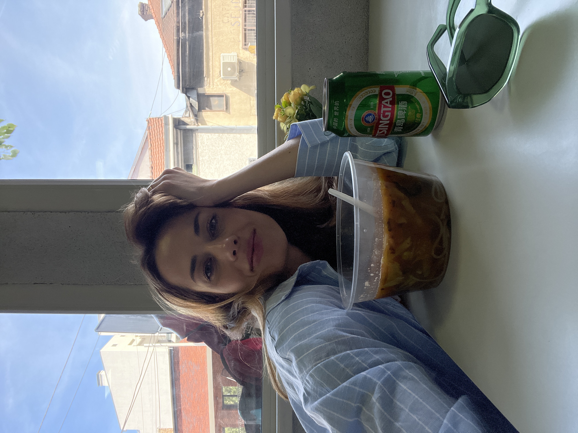 A woman is photographed indoors, likely in Belgrade, Serbia, given the architecture visible through the window. She is wearing a light blue striped shirt and leaning on her left hand. Beside her, on what appears to be a white surface, is a can of Tsingtao beer, a clear plastic container holding a liquid, possibly soup or noodles, with a straw, a small bouquet of yellow flowers, and a pair of sunglasses. Through the window, buildings with red tile roofs and light-colored facades are visible under a bright sky. A cafe sign is visible in the background.