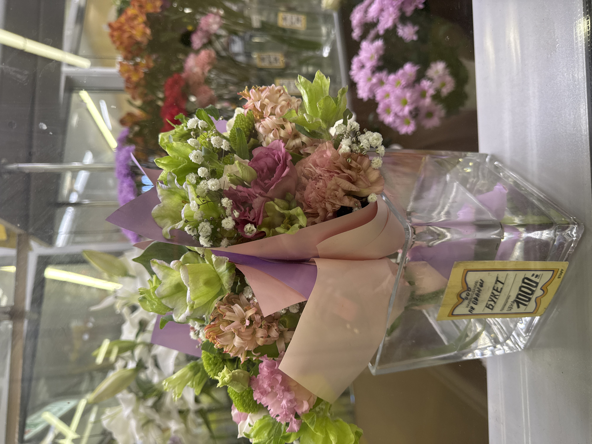 This image captures a vibrant bouquet of fresh flowers, likely displayed for sale in a flower shop or retail setting in Saint Petersburg, Russia. The bouquet is the central subject, presented upright in a clear, angular glass or acrylic container.
The floral arrangement features a mix of colors and textures. Prominent flowers include deep pink and purple roses or similar large blooms, complemented by soft peach-colored carnations. Light green, ruffled flowers or foliage, possibly green carnations or dianthus, add a fresh contrast. Small white flowers, characteristic of Baby's Breath (Gypsophila), are interspersed throughout. There are also clusters of pale peach or light pink bell-shaped flowers, resembling hyacinths. The bouquet is stylishly wrapped in layers of soft pink and lavender-purple paper or cellophane.
The background, visible through reflections in what appears to be a glass display case, shows blurred shapes of other colorful flowers, predominantly pinks, oranges, and purples. Reflections of interior lighting, possibly fluorescent tubes, indicate an indoor, commercial environment.
A yellow price tag is clearly visible on the side of the clear container. Written in Cyrillic script, it states "БУКЕТ" (BUKET), meaning "BOUQUET," and "ЦЕНА" (TSENA), meaning "PRICE," followed by "1000." This indicates the bouquet costs 1000 Russian Rubles. Other partially visible text includes "НАИМЕНОВАНИЕ" (NAIMENOVANIE - "Description") and "ОРХИДЕИ" (ORKHIDEI - "Orchids"), which might be a generic tag or refer to other items, as orchids are not overtly present in this specific bouquet. The word "STAFF" is also visible in English on the far right of the tag.
The overall impression is one of a freshly prepared and attractively presented floral gift or decoration.