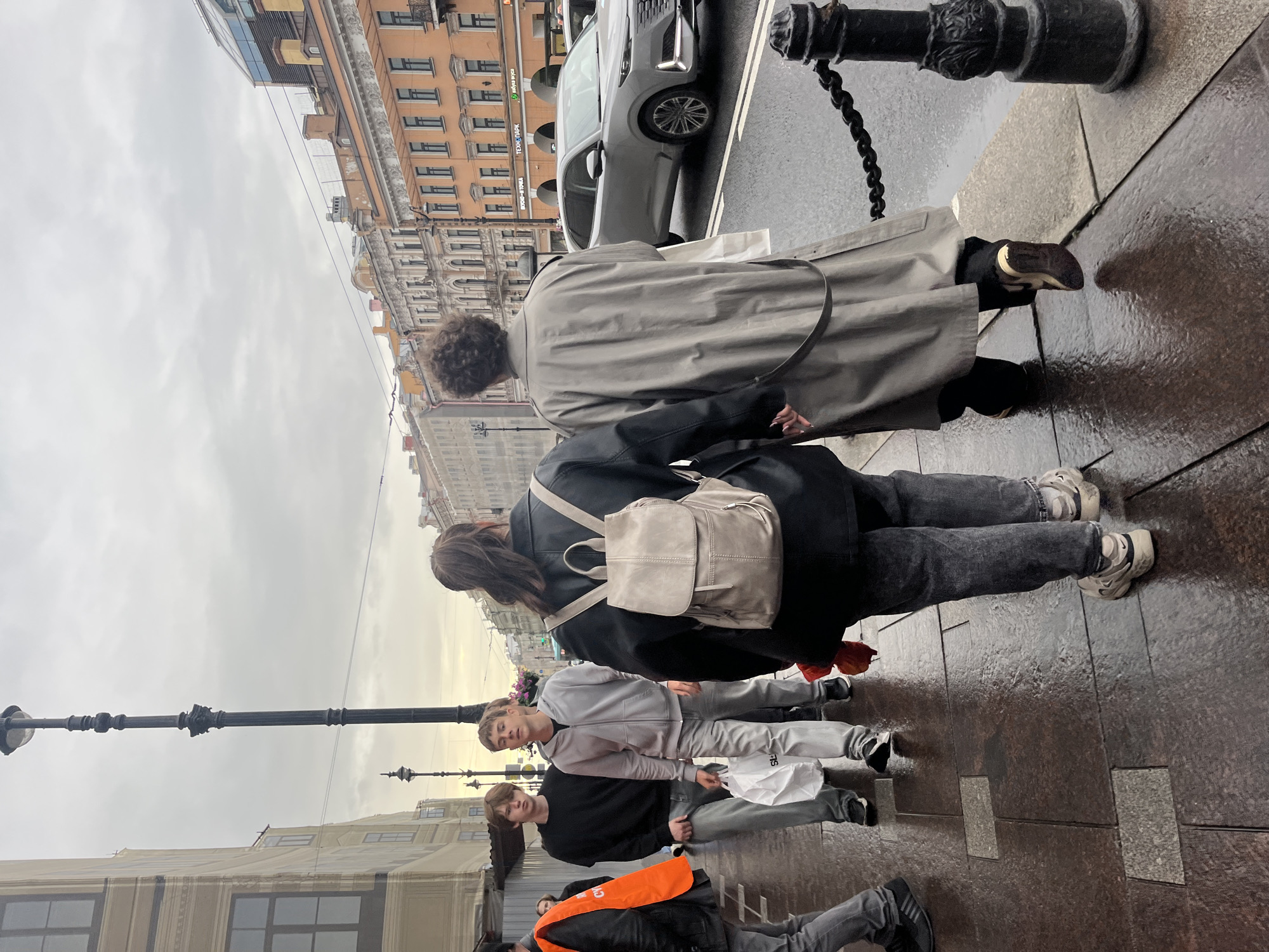 The image depicts a street scene in Saint Petersburg, Russia, on an overcast day with wet pavement, suggesting recent rain. Two people are walking away from the camera, holding hands. The person in the foreground on the left wears a black leather jacket and a grey backpack, with dark pants and light-colored sneakers. The person on the right is in a long grey trench coat over dark pants and tan and white sneakers. Other individuals are visible walking in the background, including two young men, one in a black sweatshirt and grey pants, and another in a grey hooded jacket and grey pants, carrying a white shopping bag. An orange vest is visible on a person further to the left. The background features typical St. Petersburg architecture with multi-story buildings, some with ornate facades, and a street lamp. A modern grey car is parked on the street to the right, with a black metal chain fence and bollards along the sidewalk.