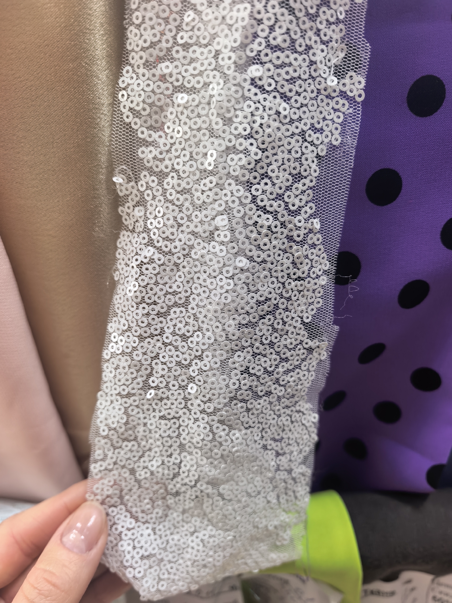 The image depicts various fabrics of different colors and textures. Starting from the left, there's a light pink fabric, followed by a shiny bronze-colored material. Overlapping these is a semi-transparent fabric covered in small, white sequins. To the right, there's a purple fabric with large black polka dots. The image appears to be a close-up shot of fabric samples or materials, possibly in a shop or crafting environment. The location, Sterlitamak, Russia, cannot be verified from the image alone. The only person appearing is a woman holding part of the sequence fabric.