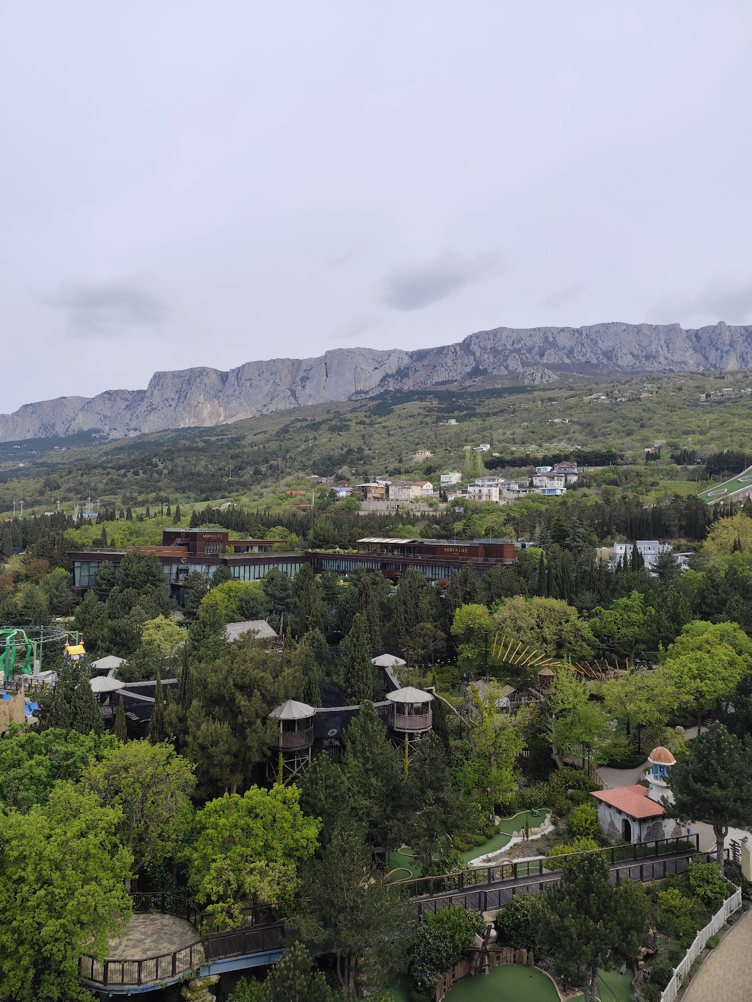 The image presents an elevated, panoramic view of a large resort complex nestled amidst lush greenery, with a prominent mountain range in the background under an overcast sky.
In the far background, a long, rugged mountain range, characterized by steep, rocky cliffs and gentler, forested slopes, dominates the horizon. Below the mountain peaks, several light-colored residential or villa-style buildings are dotted across the green hillsides.
The mid-ground features a substantial, modern building with a distinctive architectural style, including large glass windows and dark-colored exterior panels. The text "MRIYA LIFE" is clearly visible on its roofline, and "MRIYA" is partially visible on another section, indicating the name of the establishment, likely the Mriya Resort & SPA. This central building is surrounded by a dense canopy of trees, comprising both tall, dark conifers and brighter green deciduous trees.
The foreground showcases a recreational area within the resort. Multiple mini-golf courses with distinct green fairways and bunkers are discernible, along with winding pathways and wooden bridges that connect different sections. Several elevated structures resembling treehouses or gazebos with pointed roofs are interspersed among the trees. On the far left, a section of a green amusement ride, possibly a roller coaster, is visible. Another small building with a red-tiled roof and a light-colored dome is located in the lower right, adjacent to a white picket fence that runs along the frame's edge.
The scene conveys a sense of serene recreation, with the resort harmoniously integrated into its natural, mountainous surroundings. The weather appears cloudy or overcast, given the soft, diffused lighting and lack of direct shadows. No explicit human activity is captured, but the presence of various amenities suggests leisure and entertainment.