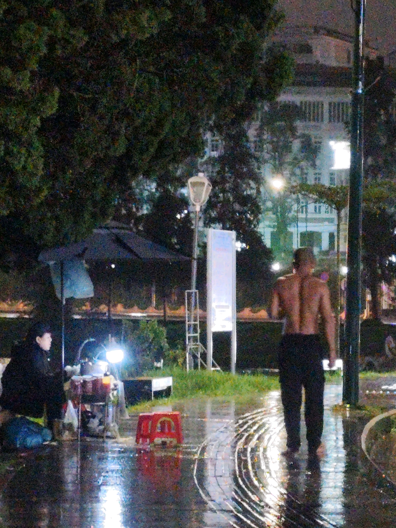 In Da Lat, Vietnam, at night, a person wearing a black jacket and hat is seated near a table with a bright lamp illuminating a collection of items, possibly for sale. To their right, a bare-chested man in dark trousers walks away from the camera on a wet, reflective pathway. The pathway appears to be paved with textured stones and is surrounded by grass and trees. In the background, a multi-story building with arched windows is visible, and streetlights cast a glow on the scene. An umbrella stands open near the seated person. The overall atmosphere is dimly lit and wet, suggesting recent rain.