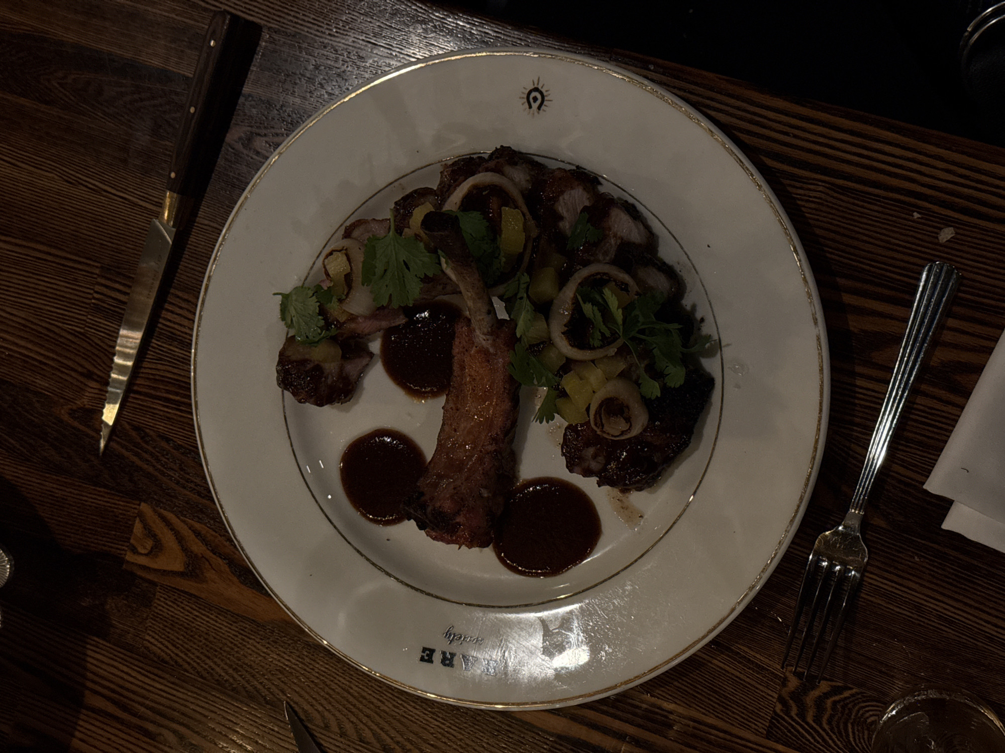 Here's a description of the image:
A top-down shot displays a culinary scene on a dark wooden surface. A white plate with a gold rim dominates the view, containing what appears to be a meat dish with a dark sauce, herbs, and some type of ring-shaped accompaniment. A knife is positioned to the left of the plate, and a fork to the right. A white napkin is partially visible near the fork. The plate bears the word "RARE" along the rim.
Based on the "RARE" branding visible on the plate, it's highly probable this photo was taken at the restaurant "Rare Society," a steakhouse with multiple locations in San Diego, CA. The specific dish and ambiance suggest a dining experience at one of their locations.