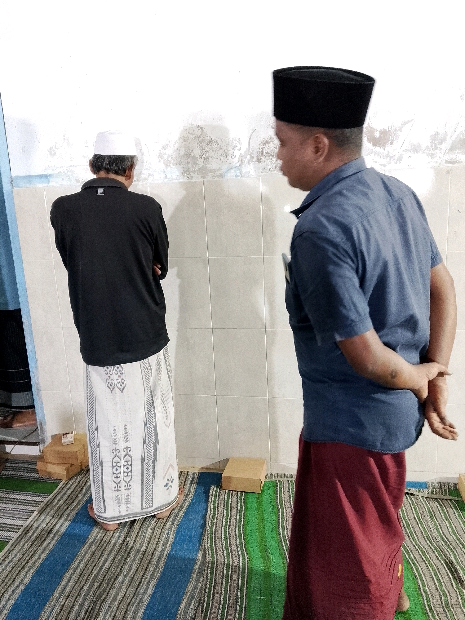 Two Muslim men are standing facing away from the camera in what appears to be a prayer room in Karangsari, Indonesia. The man on the left wears a white skullcap, a black long-sleeved shirt with a white logo on the back, and a white sarong with a patterned design. He is standing on a striped prayer mat and has his arms crossed. The man on the right wears a black peci (a cap worn by Muslim men), a blue short-sleeved shirt, and a maroon sarong. He is also standing on a prayer mat and has his hands clasped behind his back. Both men are positioned in front of a wall tiled with off-white squares. Several cardboard boxes are visible on the floor around them. The lighting suggests an indoor setting.