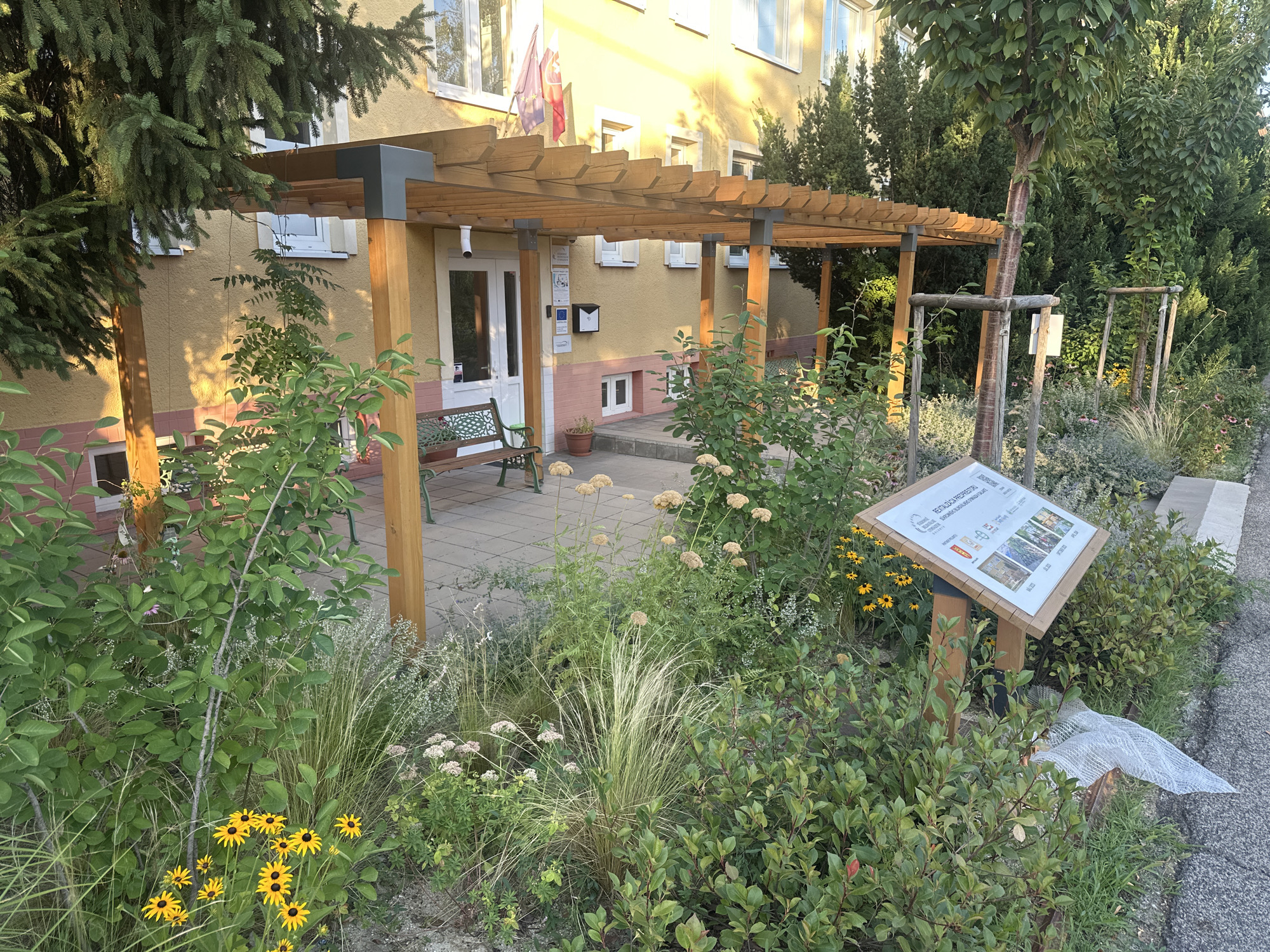 The image depicts the exterior of a building in Galanta, Slovakia, featuring a modern wooden pergola extending from the entrance. The building has a yellowish facade with multiple windows and flags displayed above the entrance. In front of the building, there is a landscaped area with various plants, including flowers, grasses, and shrubs. A metal bench sits on a paved walkway beneath the pergola. A wooden post with an informational sign is positioned in the landscaped area, displaying text and images. The overall scene suggests an outdoor space that is well-maintained and potentially serves a public function.