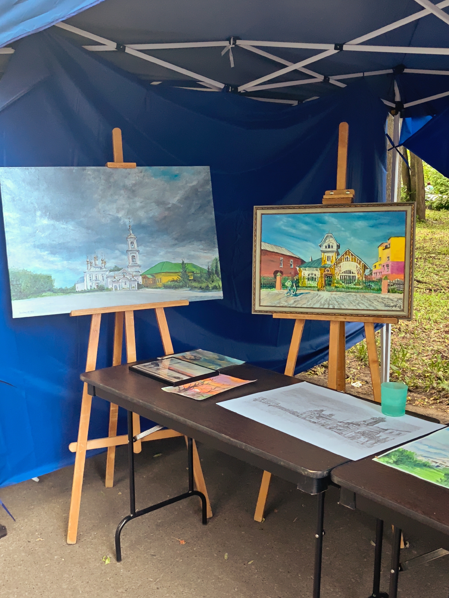 The image displays an outdoor art exhibition, likely in Kimry, Russia, given the context of the Kimry Exhibition. Two paintings are prominently displayed on wooden easels under a blue tent. The painting on the left depicts a white church with multiple domes and a bell tower, set against a dramatic, cloudy sky and a yellow building. The painting on the right is a vibrant depiction of colorful buildings with a bright blue sky, featuring two figures walking in the foreground.
In front of the easels, a dark folding table holds several artworks, including prints or sketches of architectural scenes and a watercolor painting. A green glass is also visible on the table. The ground appears to be paved, with some scattered leaves and debris. The background shows greenery and trees, suggesting a park or outdoor public space. The overall scene suggests a local art fair or market.