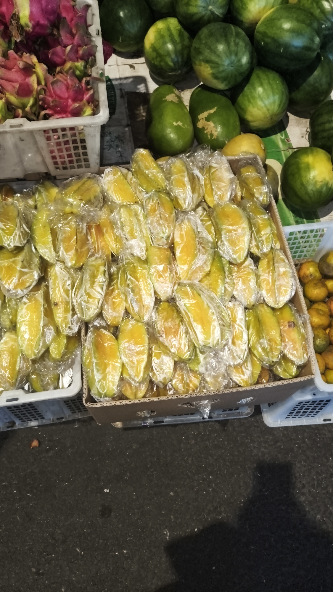 A street-level fruit stand in South Jakarta, Indonesia, displays a variety of fresh produce. In the foreground, a cardboard box is filled with star fruits, each individually wrapped in clear plastic. To the left of the star fruits, a white plastic crate holds pink dragon fruits. Behind the star fruits, a collection of watermelons and other green fruits are visible. To the right of the star fruits, a white plastic crate contains small, round orange fruits, possibly mandarins or kumquats. The ground beneath the fruit stand is dark and appears to be asphalt, with some scattered debris. The overall scene captures the vibrant market atmosphere of a local Indonesian fruit vendor.