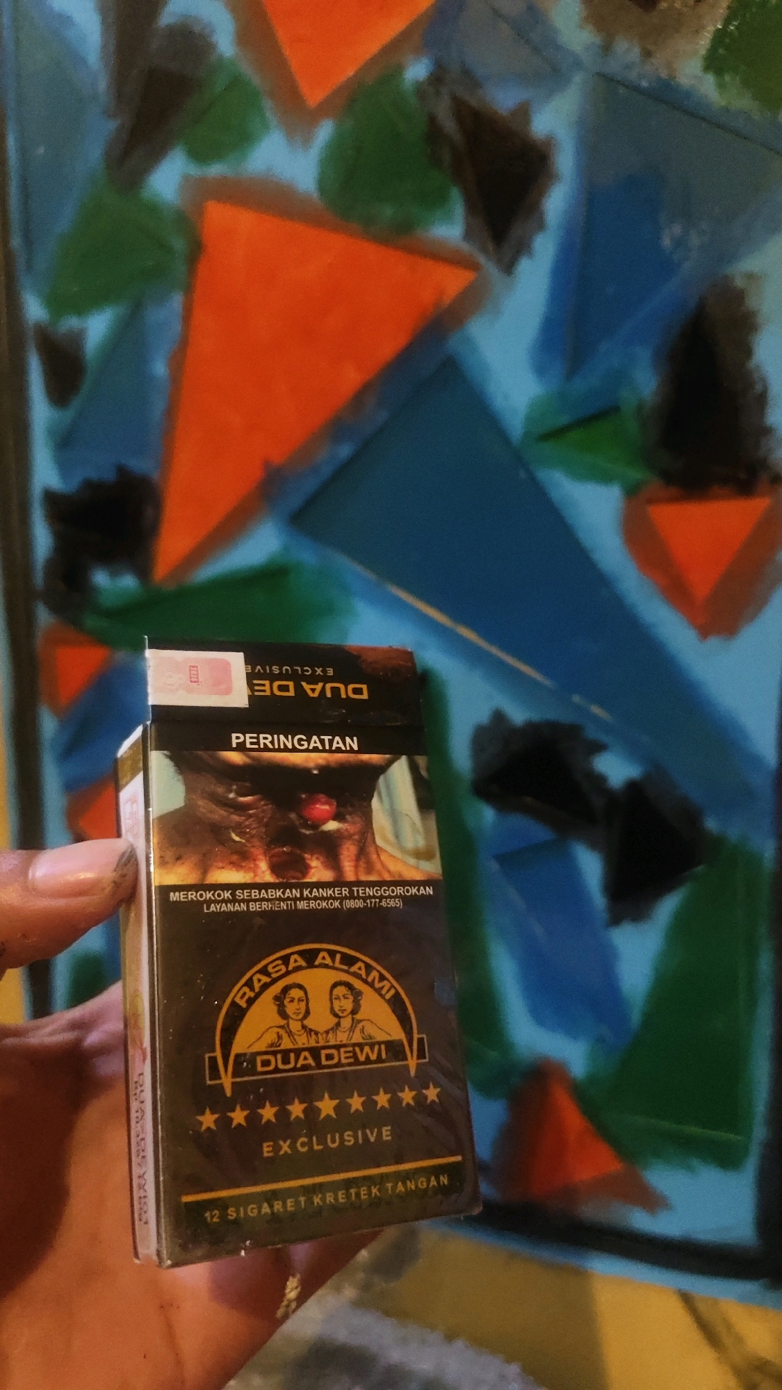 The image shows a hand holding a pack of "Dua Dewi Exclusive" kretek cigarettes. The cigarette pack features a warning label with a graphic image of a person's throat, along with text in Indonesian that translates to "Smoking causes throat cancer. Stop smoking service (0800-177-6565)". The brand logo "Rasa Alami Dua Dewi" with stars and the word "EXCLUSIVE" are also visible. The background is a colorful abstract painting with geometric shapes in shades of blue, orange, green, and black. The hand appears to be of a person of Indonesian descent, given the context of the cigarette brand and language used. There is no identifiable location in the city of Kediri, Indonesia, visible in the image, as the background is an abstract painting.