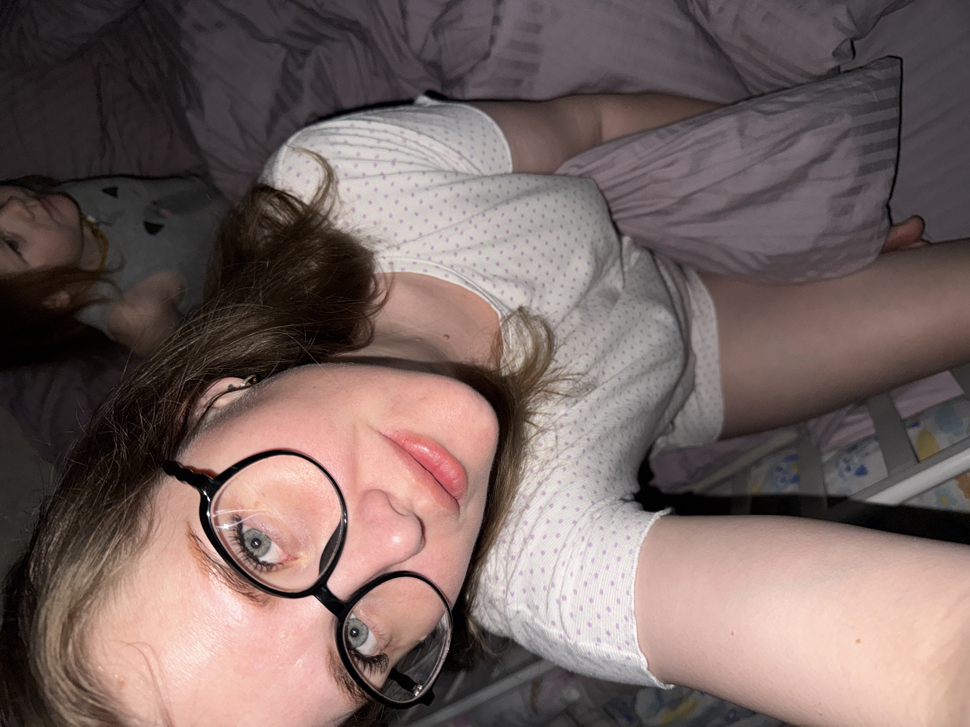 A woman wearing black-rimmed glasses and a white shirt with lavender polka dots is lying on her back in a bed with a light purple comforter. She is looking directly at the camera with her head tilted slightly. A child with reddish hair is lying beside her, also in the bed, and wearing a gray shirt with a cartoon image. The bed has white and gray bars, suggesting it may be a crib or toddler bed. The scene is dimly lit, suggesting it is night. There is no indication of the location within Gornyi (Dvurechenskoe S/P), Russia.