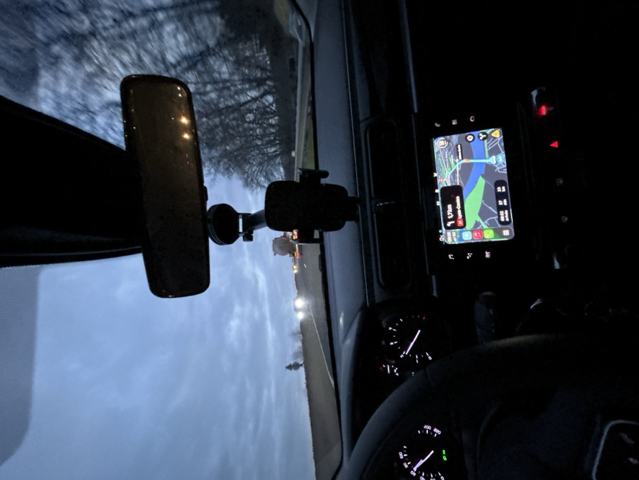 From inside a car, a view of the dashboard and front window shows a rearview mirror, phone mount, navigation screen with a map, and gauges. Outside, a twilight or early morning sky with trees is visible. Headlights from oncoming traffic illuminate the scene. The dashboard and steering wheel are only partially visible due to low lighting.