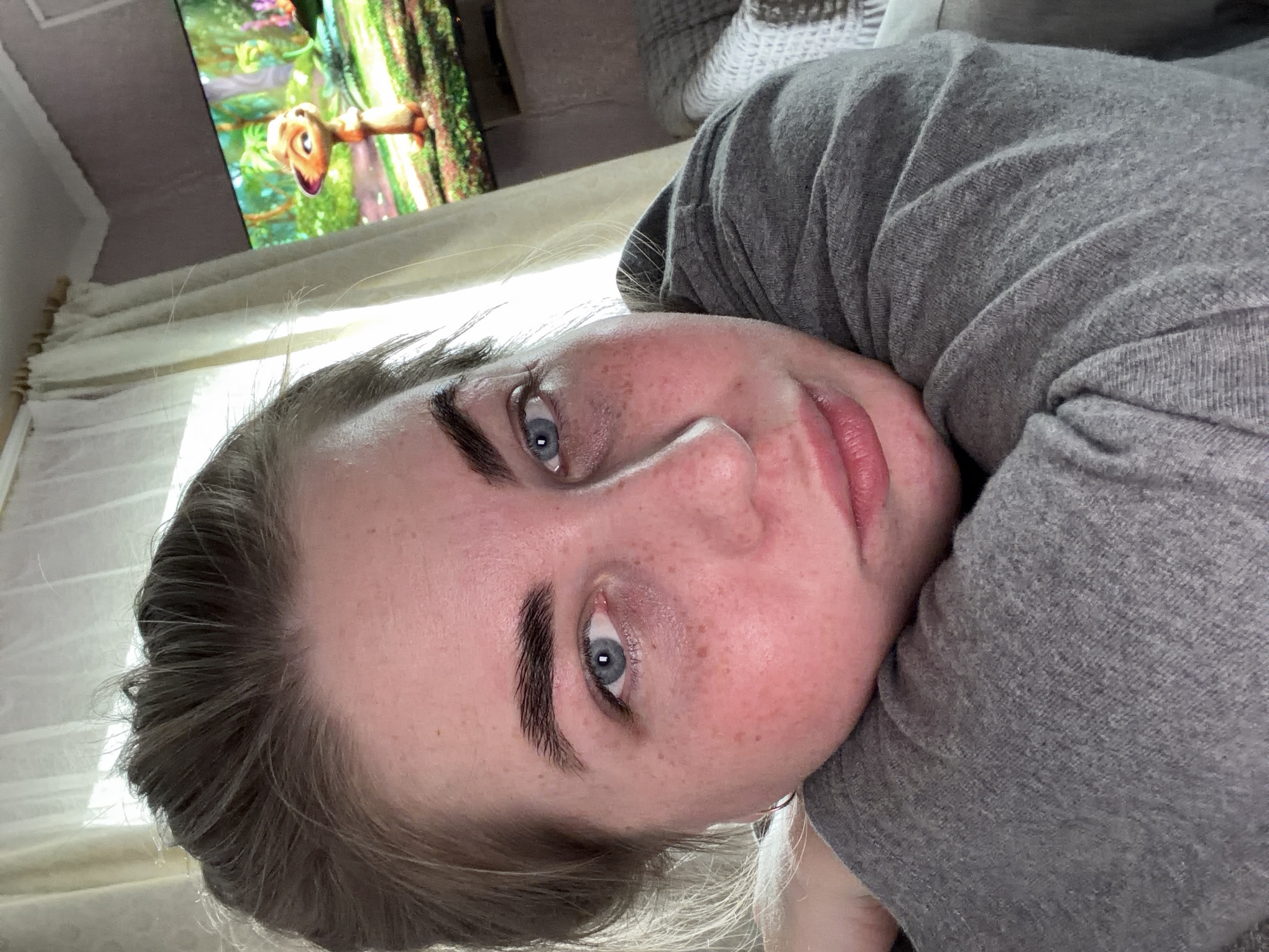 The media file is a close-up, tilted, vertical selfie of a young person lying down and looking at the camera. The subject has fair skin with freckles, blue eyes, and dark eyebrows. They are wearing a grey hooded sweatshirt. The background shows a television screen displaying a cartoon with a brown creature and a green, leafy environment, suggesting the person is in a living room or bedroom. Natural light is coming in from the left side, illuminating the person's face and casting shadows. The overall mood appears relaxed and casual. There is no visible text in the image. The location context is Уральское, Russia.
