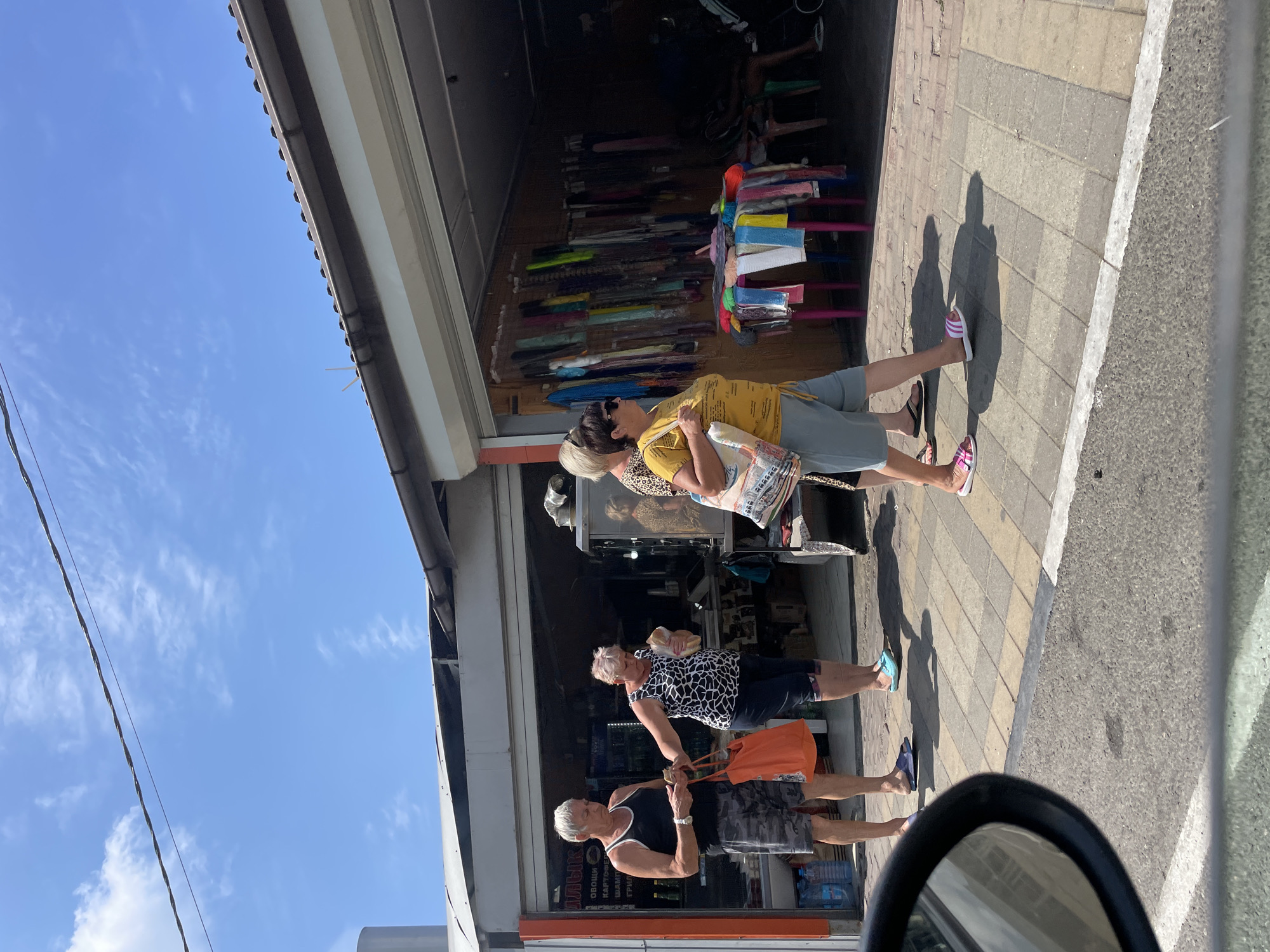 In Dzhubga, Russia, on a bright, sunny day, a scene unfolds along a roadside marketplace. Four individuals, two men and two women, are captured in a candid moment. The foremost man, clad in a black tank top and camouflage shorts, carries an orange tote bag in his left hand and appears to be holding something small in his right. To his right, a woman in a black and white patterned sleeveless top and dark shorts is extending her right hand, possibly interacting with a small white bag she holds. Behind her, a woman with short blonde hair, wearing a yellow t-shirt and light blue shorts, walks with a patterned tote bag slung over her shoulder. Another woman, with short dark hair and sunglasses, stands to the left of the yellow-shirted woman, also with a tote bag. The backdrop is a storefront featuring an open-air market with various items displayed, including colorful textiles and what appear to be food items. The ground is paved with interlocking square tiles. The overall atmosphere suggests a casual daytime outing in a commercial area.