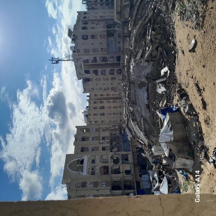 The image depicts a scene of extensive urban destruction in Khan Yunis, Palestinian Territory, under a partially cloudy, bright blue sky.
In the foreground, a sandy, disturbed ground is present, littered with various debris including small rocks and concrete fragments. Several makeshift shelters, constructed from tarpaulins and fabric in shades of white, beige, and blue, are erected on this uneven terrain.
The midground features a massive pile of rubble, consisting of pulverized concrete, twisted rebar, and other building materials, indicating a completely collapsed structure. This large mound forms a central barrier.
In the background, multiple multi-story residential or commercial buildings stand severely damaged. Their facades are heavily pockmarked with numerous bullet holes and impact craters, and many windows are shattered or entirely missing, revealing dark interiors. Sections of balconies are destroyed or missing. One building on the left shows significant structural damage on its lower floors. On the rooftop of a central-right building, a cellular communication tower or antenna array is visible, alongside what appear to be solar panels. The architectural style includes distinct arched features on some building facades.
No individuals are distinctly visible or engaged in specific actions within the frame. The overall environment suggests a landscape heavily impacted by conflict or disaster.