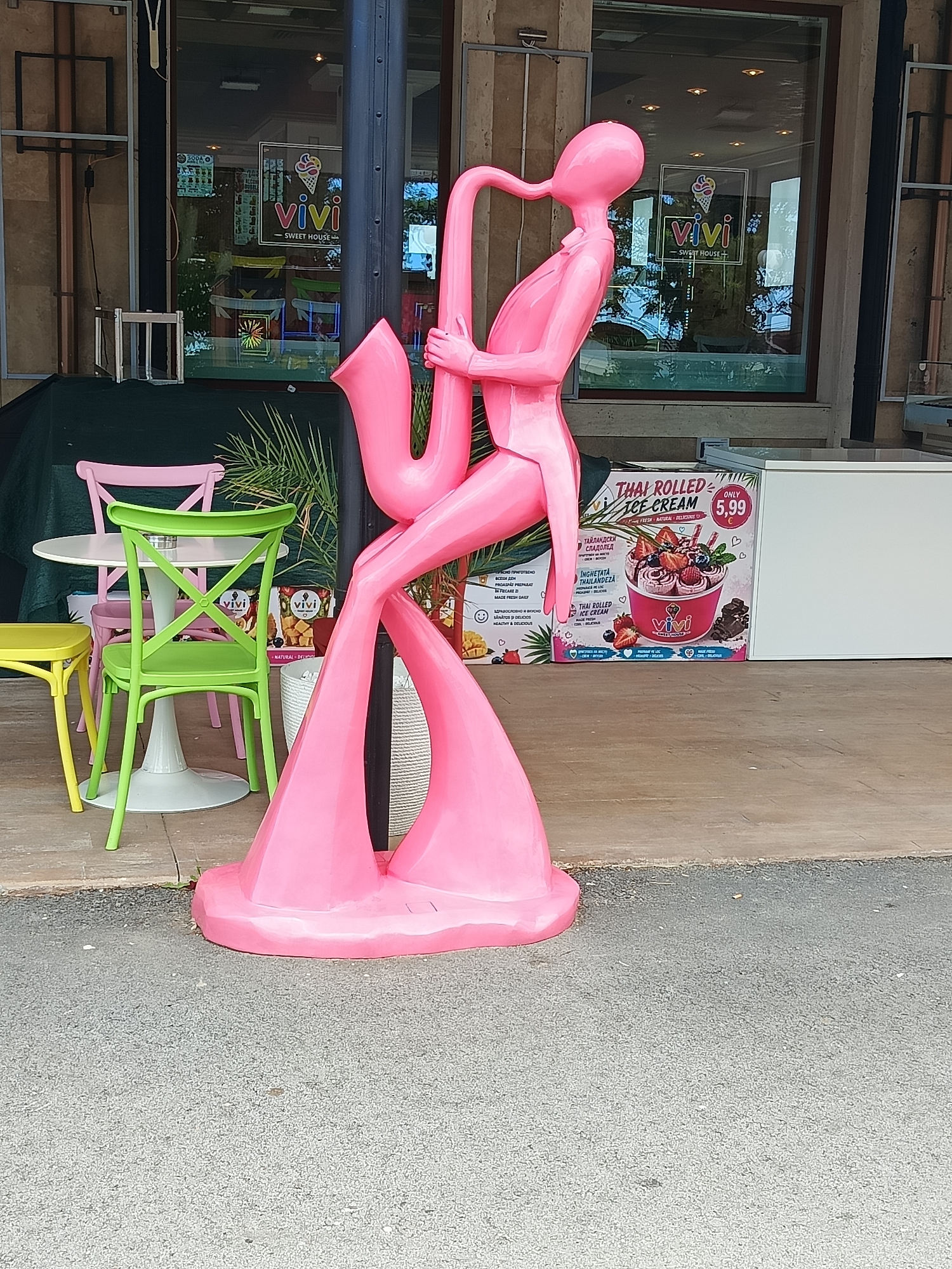 The image shows a bright pink, abstract sculpture of a figure playing a saxophone. The sculpture is life-sized or larger and is located outdoors, in front of a building with large windows that reflect the surroundings. In the background, through the windows, there is signage for "VIVI SWEET HOUSE". To the left of the sculpture, there are several brightly colored chairs (yellow, green, and pink) around a white round table. Behind the table, there is a palm frond. To the right of the sculpture, there is a sign advertising "THAI ROLLED ICE CREAM" with a price of 5.99. The ground appears to be paved, possibly a sidewalk or outdoor seating area. The overall scene suggests an outdoor seating area of a cafe or dessert shop. The location is Varna, Bulgaria, as indicated by the presence of the "VIVI SWEET HOUSE" signage, which is a known business in Varna.