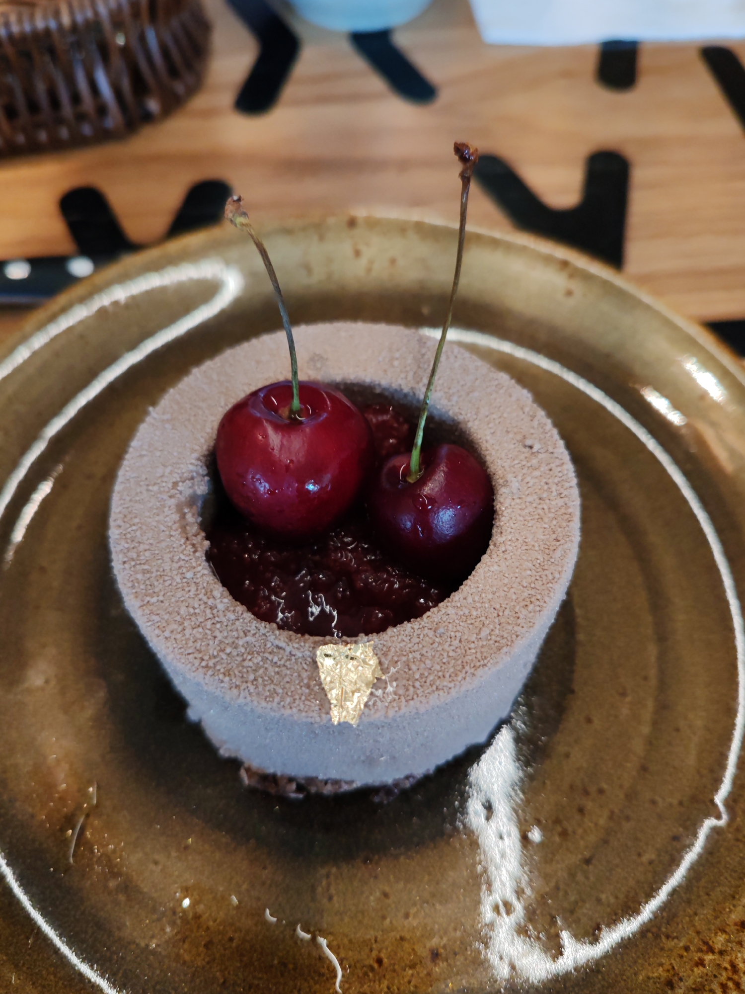 A close-up, overhead shot shows a dessert on a plate, likely in Syktyvkar, Russia. The dessert is a small, cylindrical cake with a textured, light brown outer layer, resembling velvet or a fine crumb topping. The top of the cake is hollowed out, revealing a dark red, jam-like filling. Two fresh cherries with stems are placed on top of the filling. One cherry is positioned towards the left side, and the other is slightly to its right, partially obscuring the filling. A small piece of gold leaf is adhered to the front-facing side of the cake's outer layer. The cake sits on a rustic, earthy-toned ceramic plate with a slightly raised rim. The background is blurred, but hints of a woven basket, a wooden surface with dark markings, and a hint of white suggest an indoor setting, possibly a restaurant or cafe.