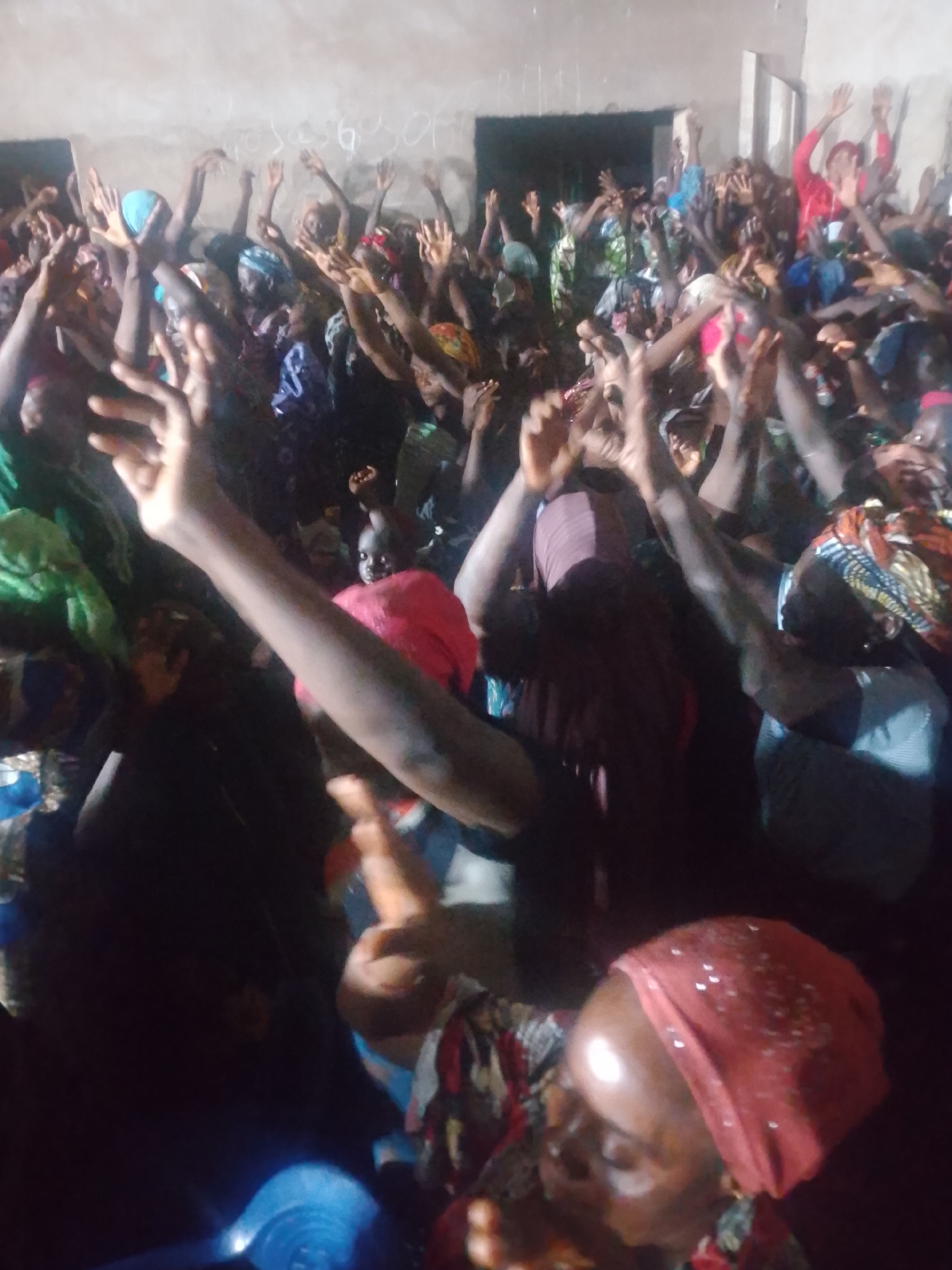 A dimly lit, crowded indoor scene depicts a group of people, predominantly women, with their arms raised in the air. The individuals are wearing colorful headwraps and clothing, suggesting a celebratory or religious gathering. The background appears to be a plain, unadorned wall, with some faint markings or writing visible. The lighting is harsh and casts shadows, with some areas appearing overexposed due to the direct light source. The overall impression is one of collective energy and participation within a community setting, possibly a church or a similar assembly.