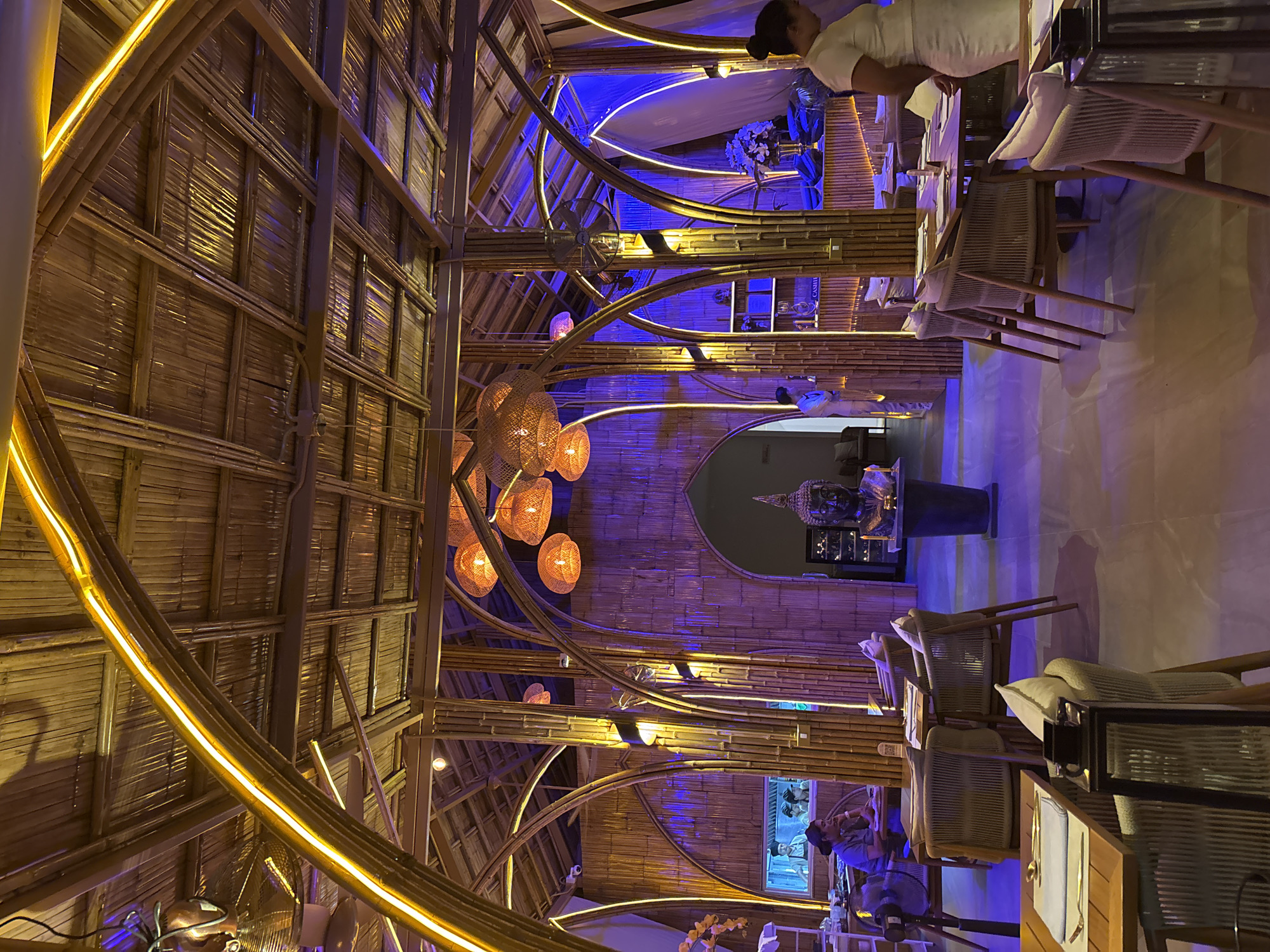 This media file captures the interior of an upscale restaurant or lounge, showcasing a unique architectural design predominantly featuring bamboo. The setting is illuminated by prominent artificial lighting, indicating an evening or nighttime ambiance.
The main subjects include numerous bamboo structures forming the ceiling, walls, and decorative archways that span the space. Warm yellow LED strip lighting is integrated into many of these bamboo beams, creating glowing accents throughout. This warm lighting is contrasted by cool blue and purple ambient lights washing over sections of the bamboo walls and the polished floor, particularly on the right side of the image.
Several spherical, woven pendant lights with a warm orange glow hang from the ceiling, adding to the exotic decor. A large, dark-colored Buddha statue, placed on a pedestal within a recessed archway on the right wall, serves as a central decorative element.
The scene depicts a dining or social event. Tables are set with white placemats and cutlery, accompanied by modern chairs with woven backs and light-colored cushions. Several people are visible: staff members dressed in white uniforms are seen near tables and in the background, while diners are seated at various tables.
Notable details include ceiling fans mounted on the bamboo structures, and a partially obscured blue-lit sign on a far wall that seems to read "CASII...". Small signs, possibly QR codes, are visible on some tables. The extensive use of bamboo, the Buddha statue, and the sophisticated lighting create an exotic, tranquil, and luxurious atmosphere, characteristic of a high-end establishment in a location like Baan Lamai, Thailand.