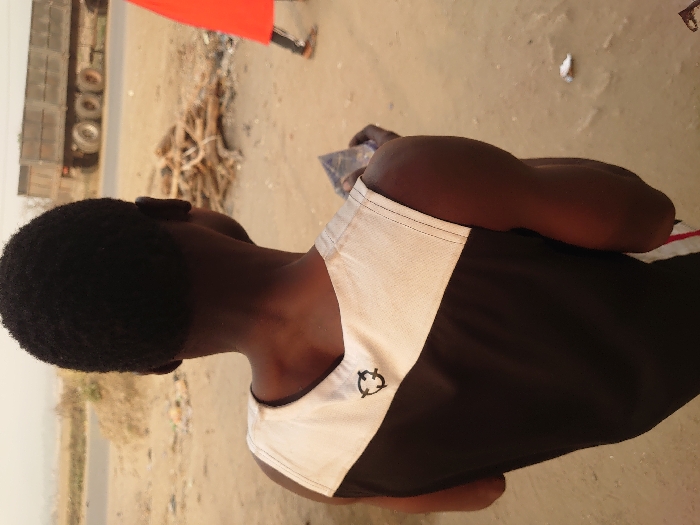 The image presents an outdoor scene in Ajaokuta, Nigeria, characterized by dry, dusty conditions. In the foreground, a person with dark skin and short, dark hair is depicted from the back, slightly over their right shoulder. This individual wears a sleeveless garment that is predominantly black, with a cream or light tan upper panel featuring a black, stylized circular emblem near the left shoulder blade. Their right arm is bent, holding an indistinct dark, crumpled object.
The person stands on uneven, light brown, dusty terrain. In the midground, a paved road extends horizontally across the frame. Adjacent to the road, a pile of stacked wooden branches or logs is visible. To the far right, a second individual's lower body is partially visible, clad in a bright red garment and brown footwear.
In the background, a large, multi-wheeled grey heavy-duty truck, possibly a dump truck, is situated on the paved road. The distant horizon appears hazy, consistent with atmospheric dust or a far-off landscape. The overall lighting suggests daylight.