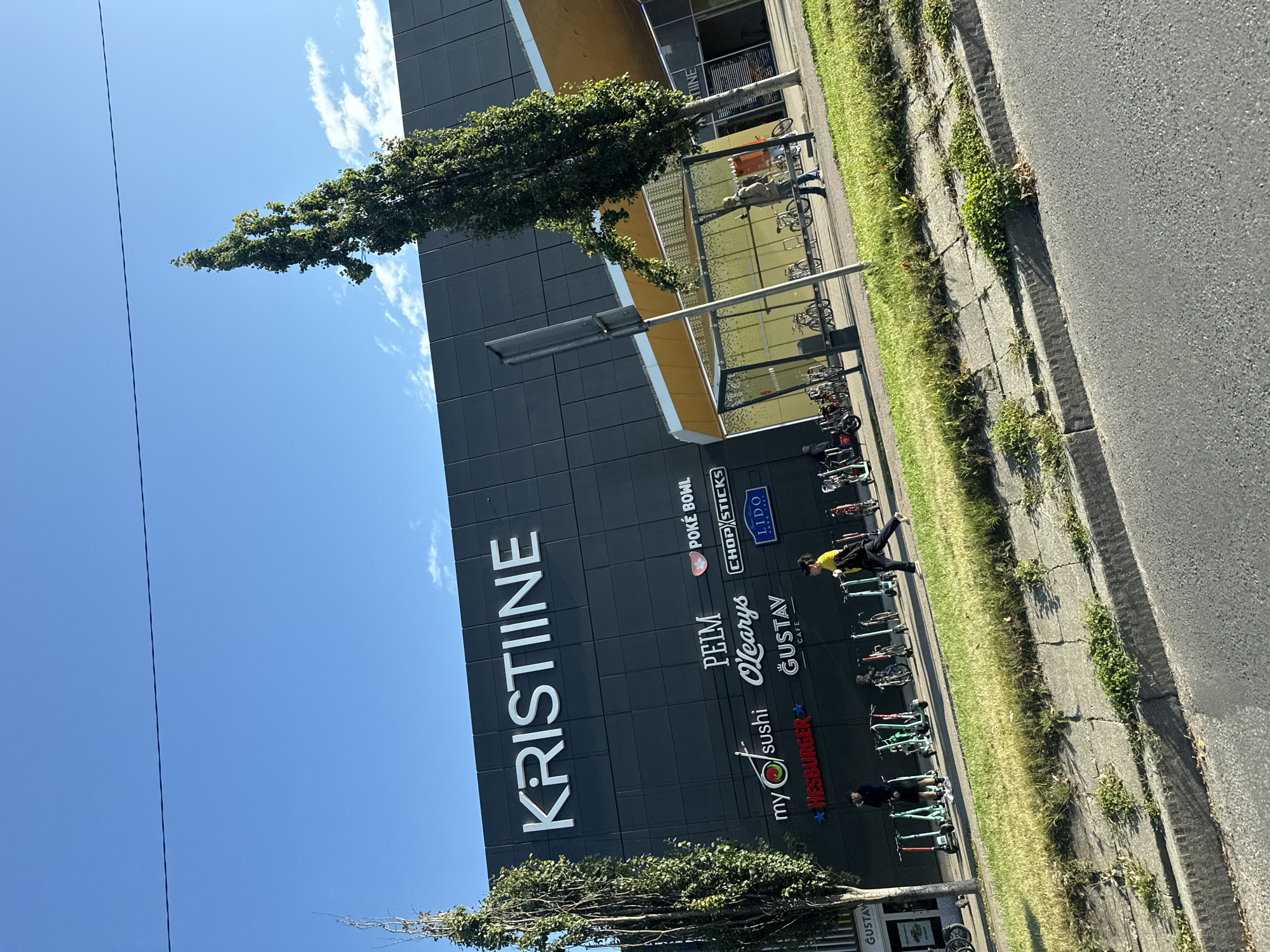 The image depicts the exterior of the Kristiinne shopping center in Tallinn, Estonia. The large, dark gray building features prominent white lettering spelling out "KRISTIINE" across its facade. Below the main signage, various restaurant and shop logos are visible, including "my sushi", "PULM O'Learys", "POKÉ BOWL", "CHOP STICKS", "LIDO", and "GUSTAV CAFE". A golden-colored canopy extends from the building, covering an area with bicycles parked underneath. Several people are present on the sidewalk in front of the building, with some walking and others standing. A line of electric scooters is parked to the left of the building. The scene is set against a clear blue sky with a few scattered clouds. The foreground shows a grassy verge and a concrete curb with an asphalt road. The overall impression is a modern urban commercial area on a sunny day.
