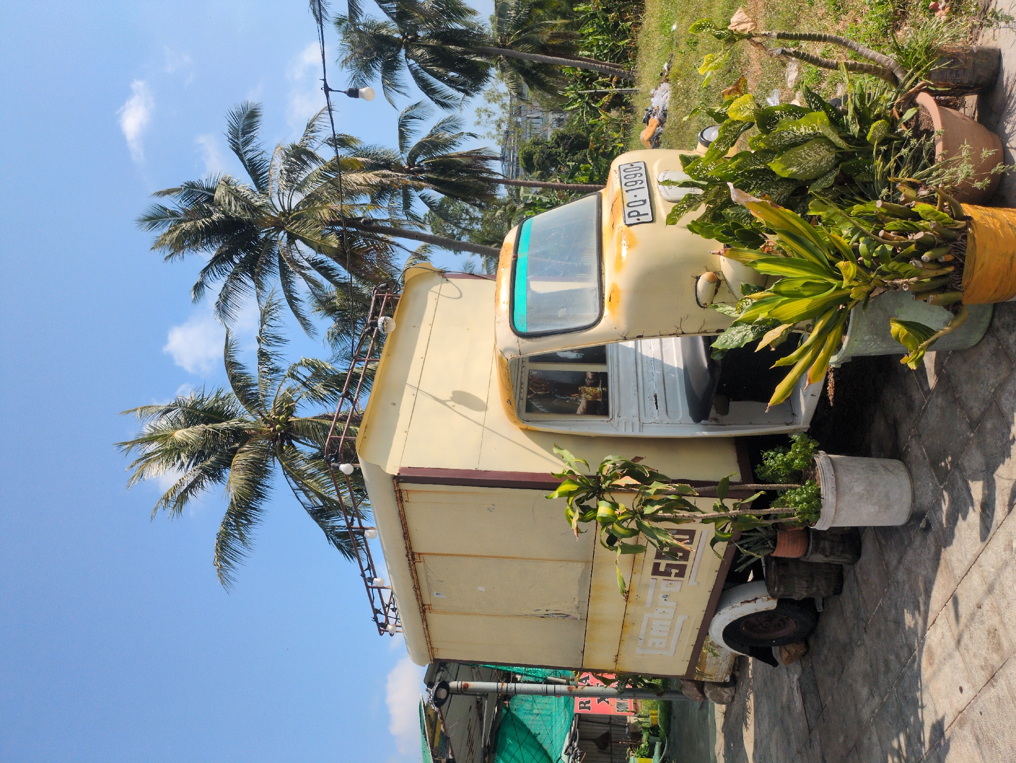 The image depicts a repurposed vehicle, possibly a van or truck, converted into a decorative display or potentially a stationary shop. The vehicle is painted in faded yellow and is located outdoors. Its front bears a license plate. Various potted plants and foliage surround the vehicle. The scene is set in a tropical or subtropical environment, evidenced by the presence of palm trees in the background against a bright sky with scattered clouds. The location is Phú Quốc, Vietnam. The surrounding environment consists of vegetation and what seems to be infrastructure, possibly a building or some sort of establishment.