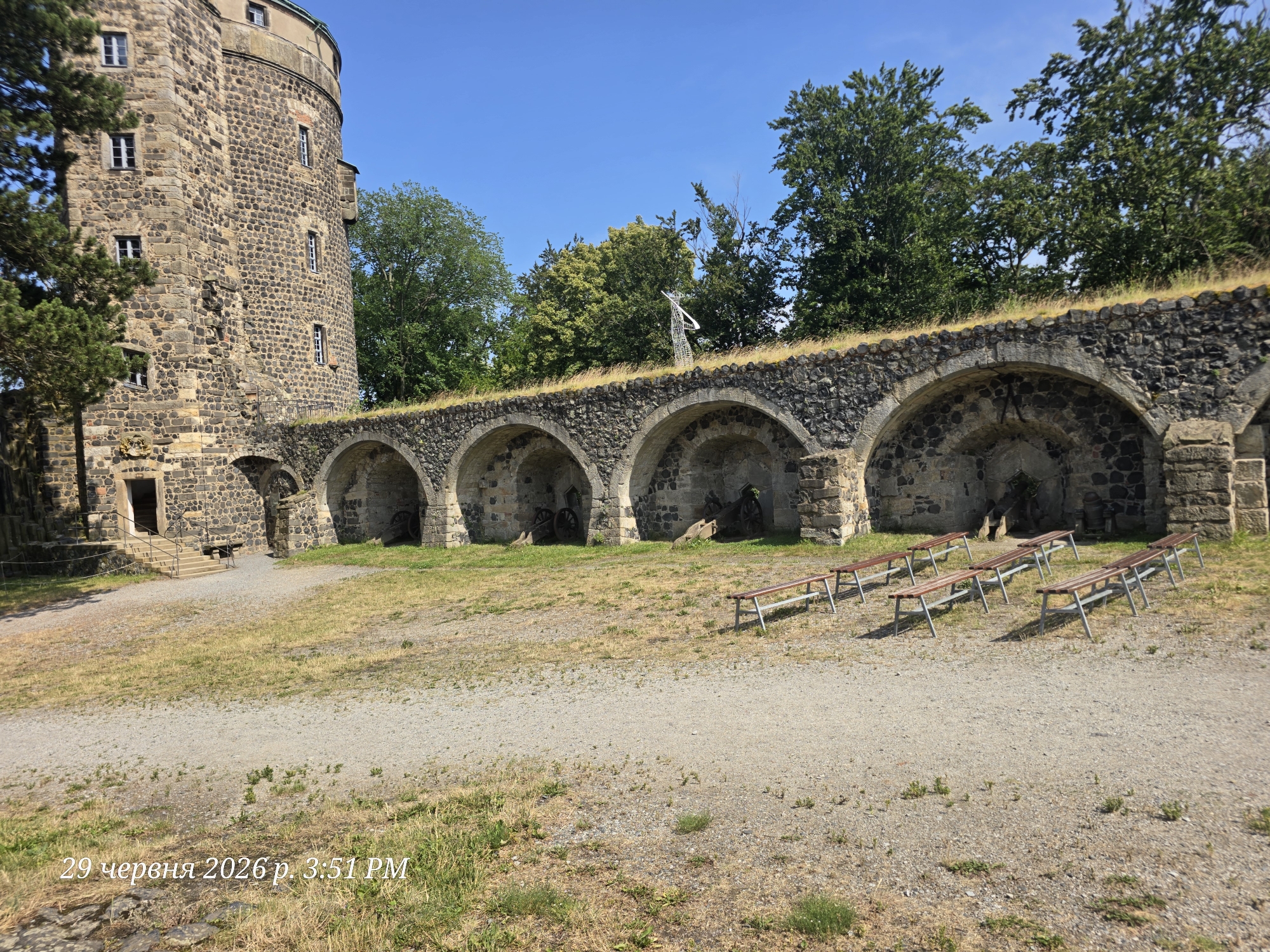 This media file captures an outdoor scene featuring a historic stone tower and an arched walkway, likely part of a castle or fortification. The tower, constructed from rough-hewn stones, has multiple small windows and a circular design. Adjacent to the tower is a long, arched structure with several openings. Inside these arches, old cannons are visible, suggesting a historical defense site. The walkway is lined with a row of wooden benches with metal legs, arranged facing outwards from the arches. The ground is covered with gravel and sparse dry grass, indicating a somewhat arid environment. Lush green trees are present in the background, providing a contrast to the stonework. The sky is clear and blue, suggesting fair weather. The lighting indicates it is daytime, likely late morning or early afternoon, given the shadows. A white, abstract sculpture is visible atop the arched structure, adding a modern element to the historical setting. The overall mood is serene and perhaps a little somber due to the presence of the cannons.
Visible text in the lower left corner reads "29 червня 2026 р. 3:51 PM", which is a date and time stamp. The location context provided is Stolpen, Germany. No people are visible in the frame.
